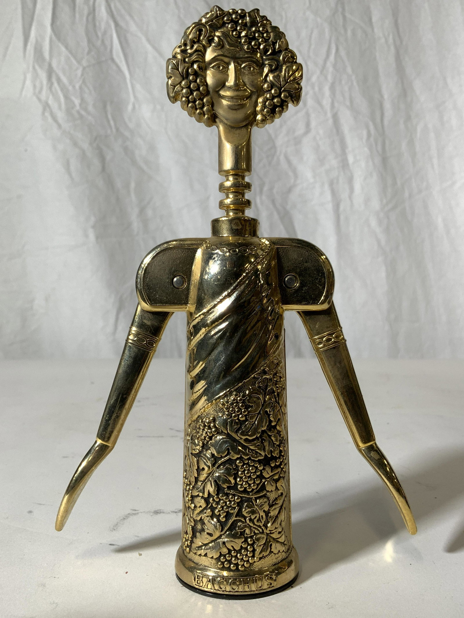 GODINER ITALY BACCHUS Corkscrew: Piece reads BACCHUS GODINGER ITALY AT BASE. Piece is gold toned, possibly brass. Corkscrew has grape, vine and leaf detail. Top of piece depicts women’s face. Piece measures approx 8.5 inches tall 2