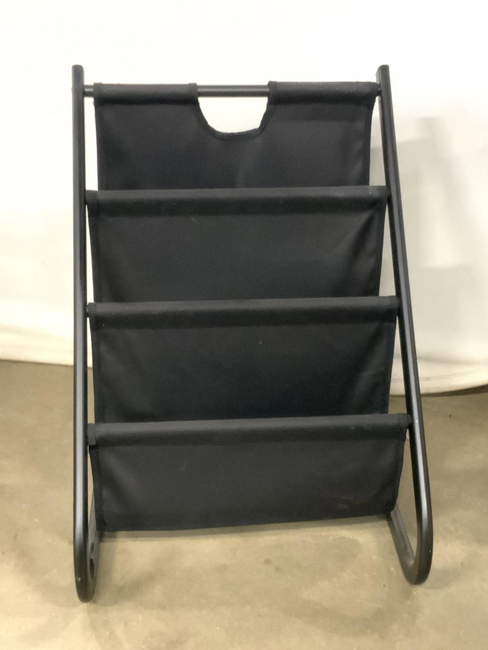 Black Metal & Fabric Magazine Rack: A magazine rack crafted from metal and fabric materials. Measures Approx. 22H x 16W in. Measured at widest point. Some surface wear. Missing piece to reverse. Magazine rack, metalware, metalwork, home