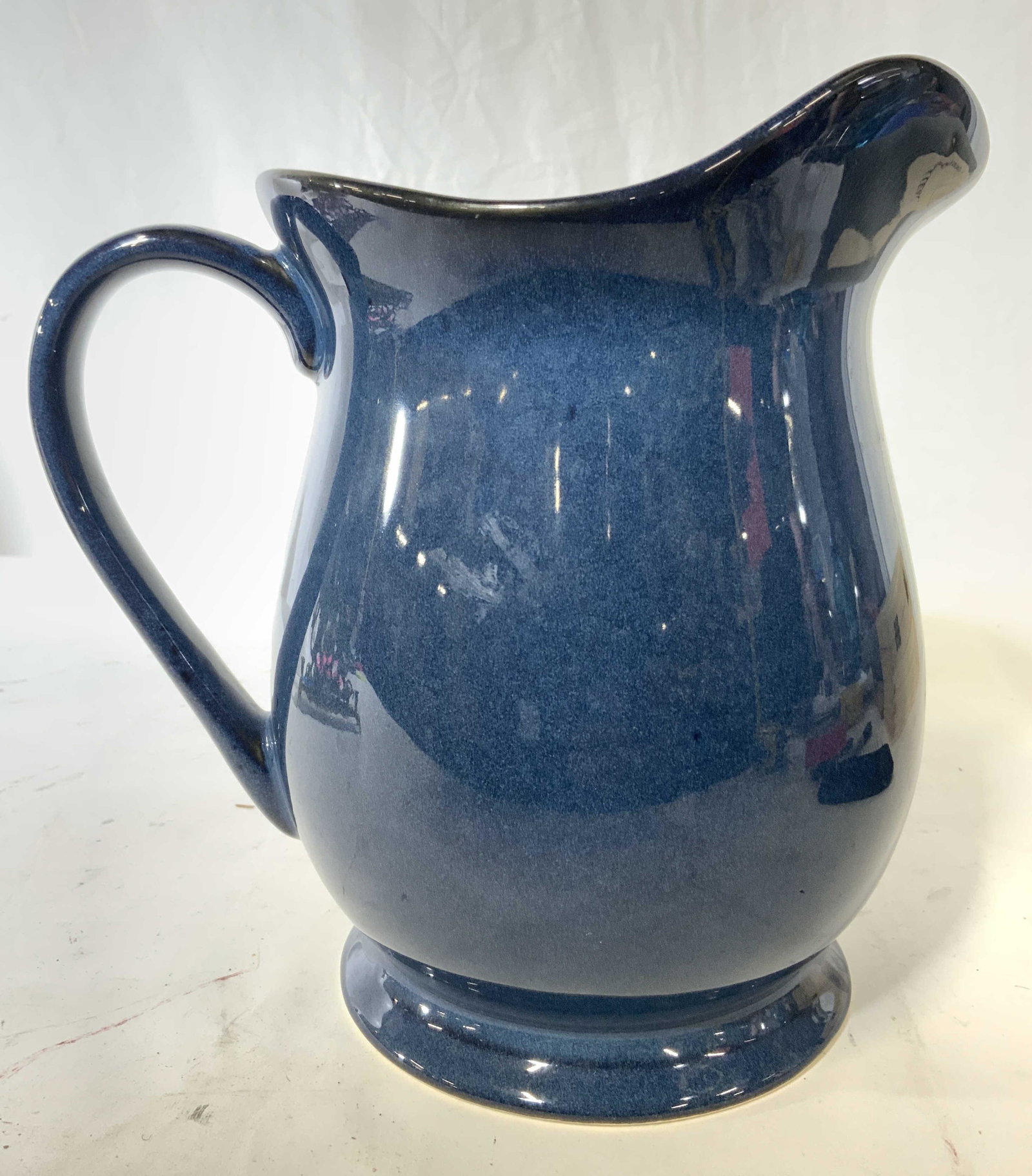 Footed Porcelain Pitcher W Glazed Finish: Pitcher appears to be porcelain possibly ceramic. Piece is blue toned and footed. Underside reads JC PENNEY. Pitcher has glazed finish. Piece measures approx 8.5 inches tall 6.5 inches wide 9 inches d
