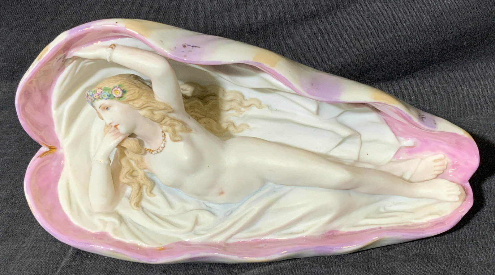 Reclined Female Nude Figurine: Reclined Female Nude Figurine, no apparent inscriptions or mark to piece, subject a ceramic reclined female nude within a shell like vessel, measures 4 x 4 x 8 inches, surface wear and crack to piece.