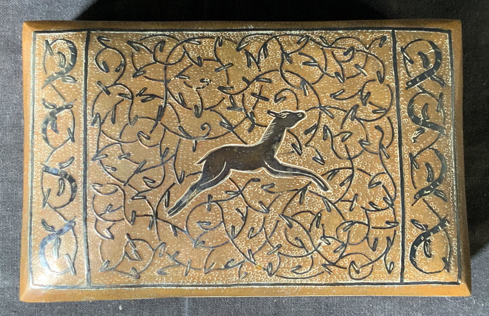Bronze VNTG Toned Metal Deer Trinket Box: A box lidded made of bronze toned metal, with black raised mythical designs of foliage and leaves. The lid has a depiction of a fawn. Interior of the box is lined with wood. Underside has 2 felt chair