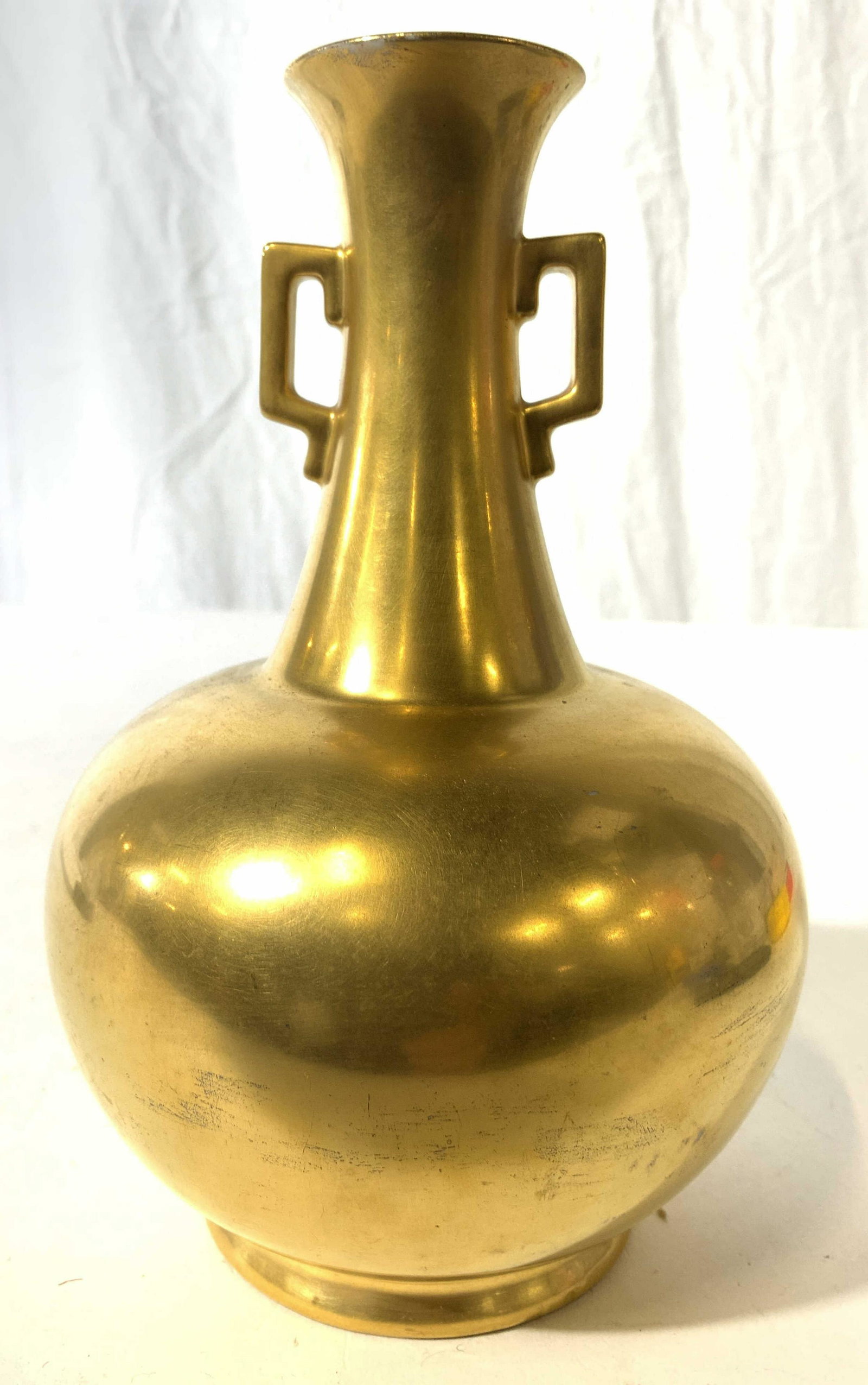 Gold Gilt Porcelain Vase W Handles: Vase is gold toned and porcelain. Piece appears to be covered in gold gilt. Piece has 2 handles on neck. Vase has narrow neck and wide seat lower body. Base of piece is footed. Piece measures approx 7