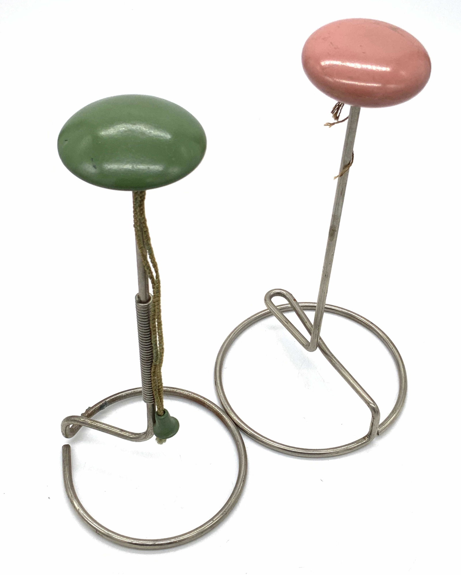 Lot 2 Vintage Metal Wig Stands: Two vintage metal wig stands. Pieces have a green and a pink topper. Similar is size. Measures approx 9H x 4.5W in. Pieces have surface wear, some scratches. Pink one has no cord. Vintage collectibles