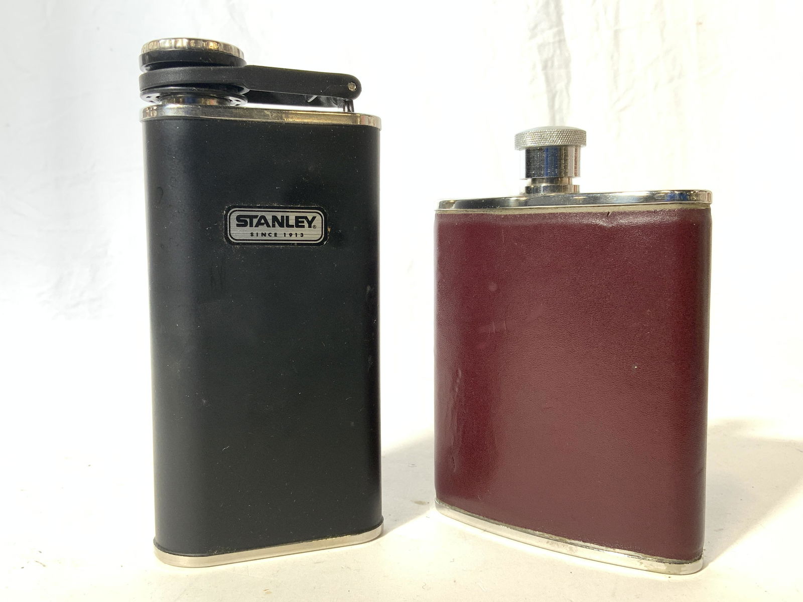 Lot 2 Metal Flasks W Lids: Lot includes 2 flasks. One black and silver toned and one maroon and silver toned. One flask reads STANLEY, One flask appears to be leather lined. Largest flask measures approx 6.5 inches tall 3.25 in