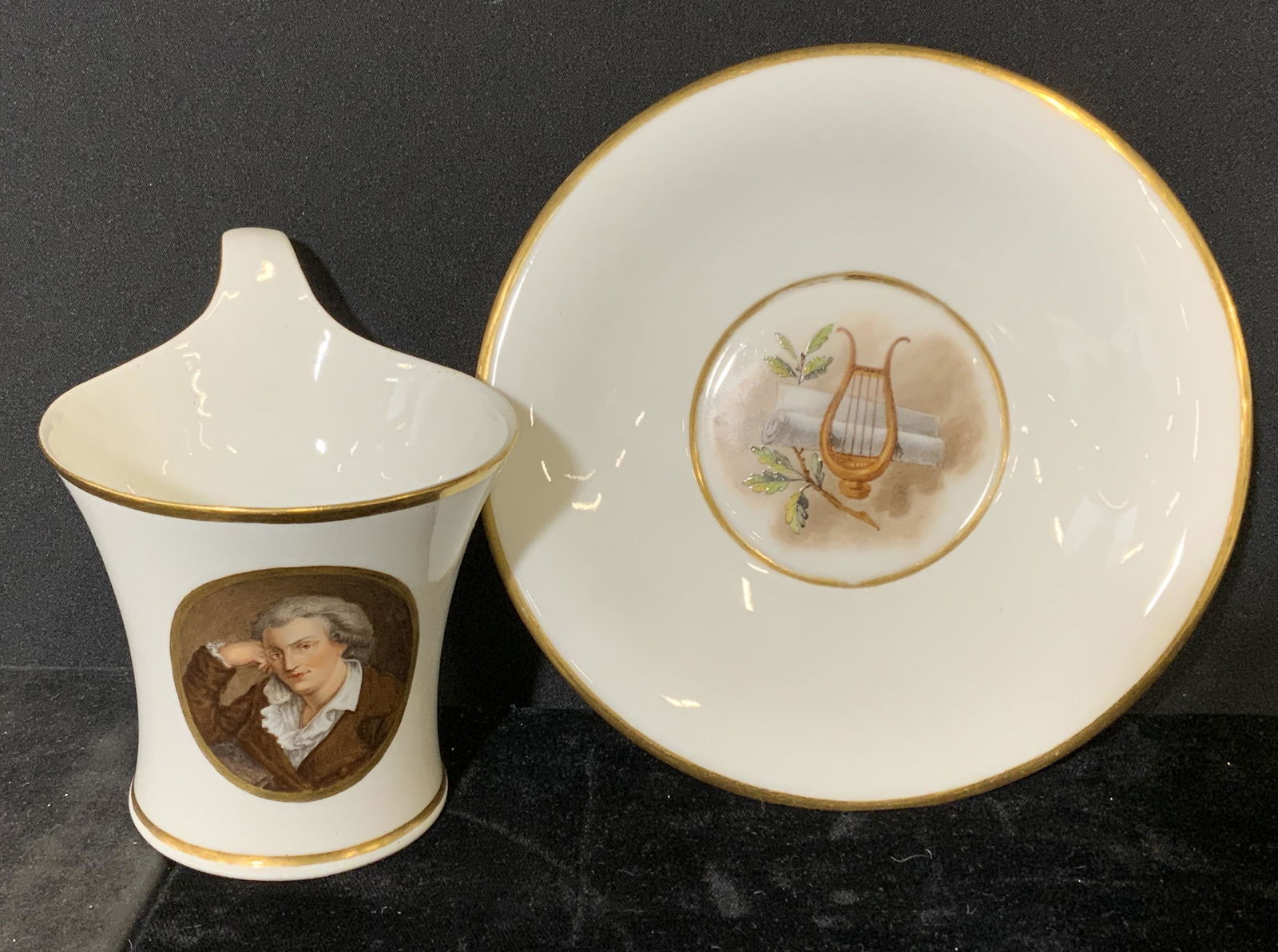 Vintage Tea Cup w Saucer: Vintage tea cup with saucer. Cup decorated with an image depicting a male figure, portrait bordered by gilt paint. Well of saucer depicts a lyre and a scroll. Wear is consistent with age and use, chip
