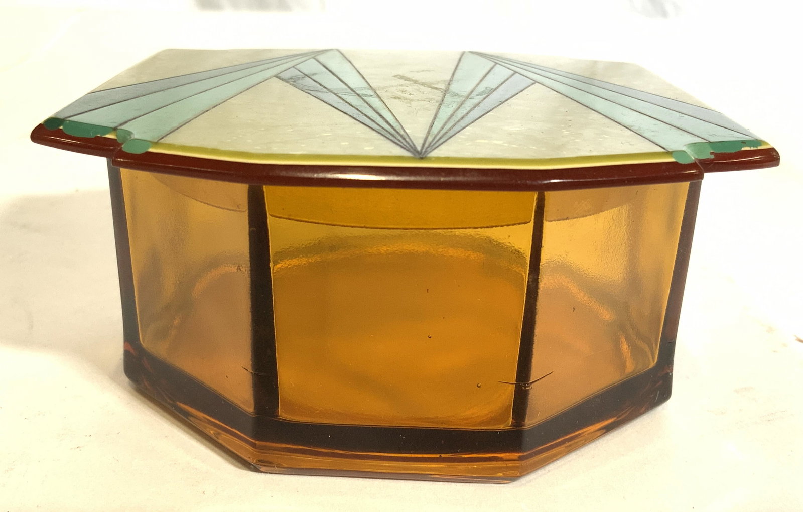 Amber Glass Trinket Box W Enameled Lid: Trinket box is Amber toned and octagonal shaped. Lid appears to be enamel, possibly Bakelite, possibly catalin. Lid has m shaped detail. Trinket box with lid measures approx 4 inches wide & deep 2 inc
