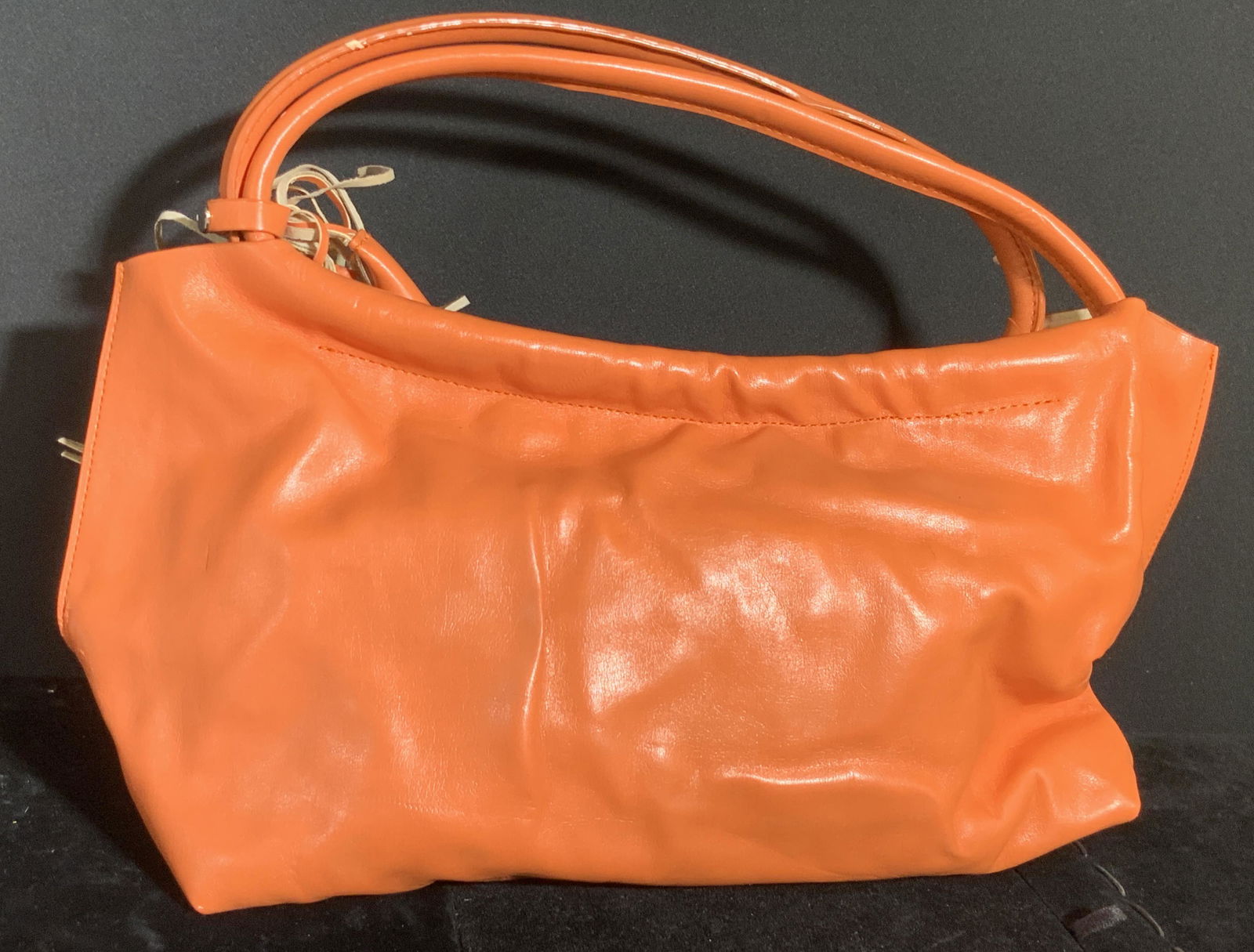 Vintage Orange Leather Style Ladies Purse: A vintage orange hued leather style ladies purse. Two top handles. Tassel accent to the side. Snap button closure. Interior is lined with suede style material. Measures approx 7L x 15W in. Indentation