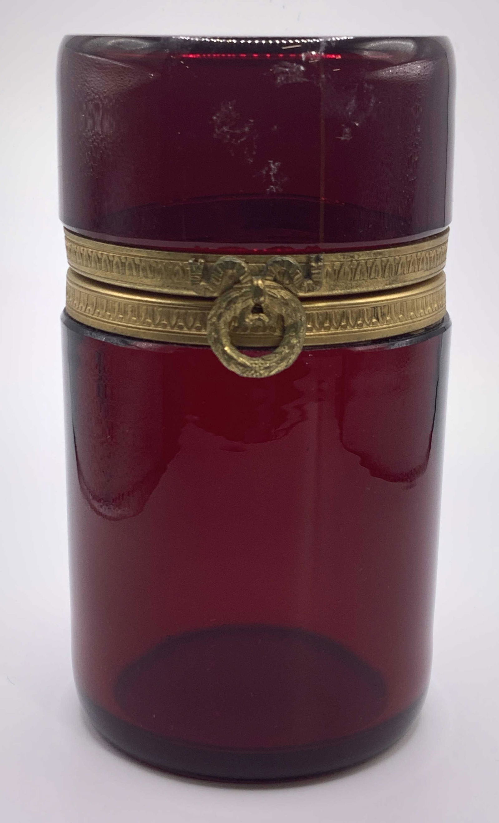 Red Toned Glass Lidded Vessel: A hinged lidded glass vessel, dresser box, with the glass being a Ruby toned red. Rim of vessel and lid are covered in a gold toned metal with an intricate raised pattern and a gold metal reef on the