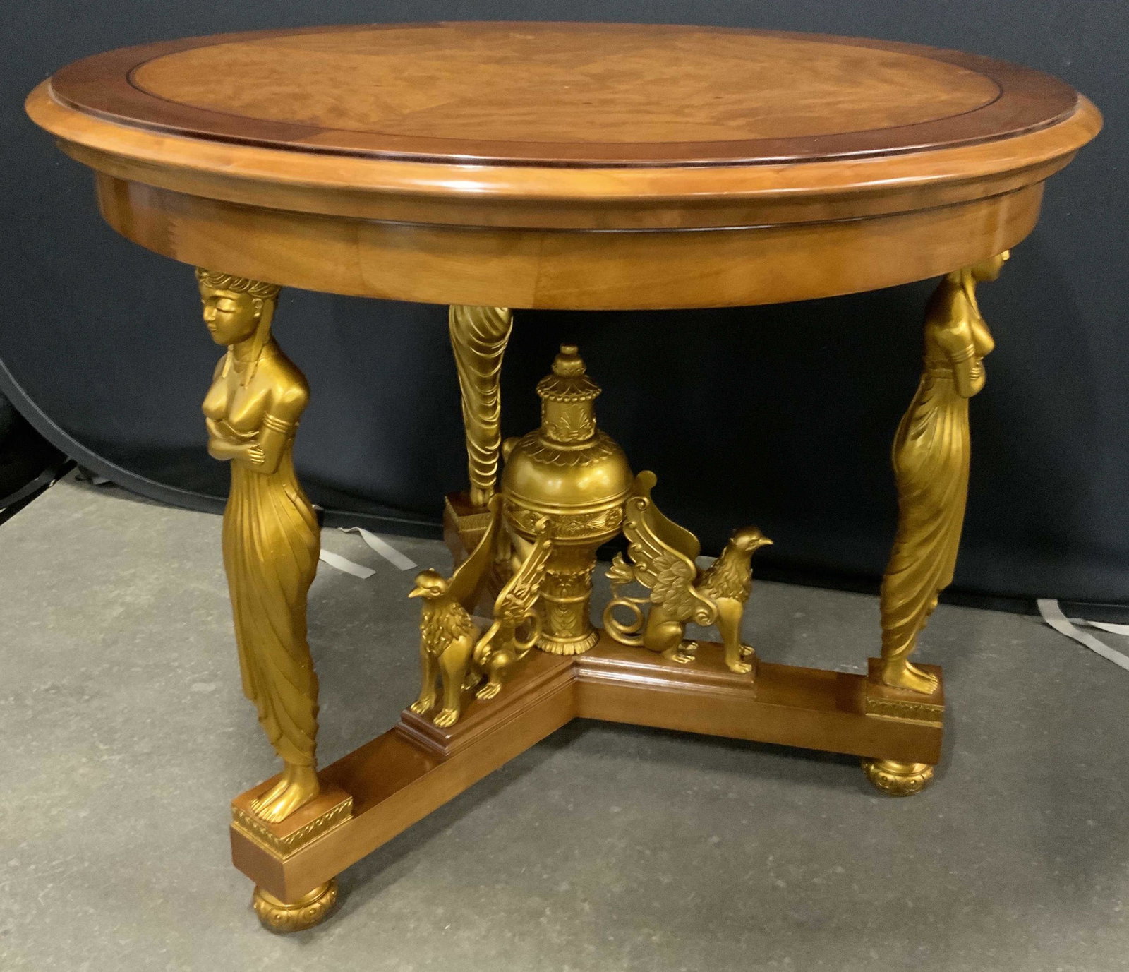 Regency Style Wooden Center Table: Wooden table, possibly vintage. Multi toned veneered surface. Gilded, carved supportive detailing. Depicts several griffins, and robed female figures, large urn like finial seats between the griffins.