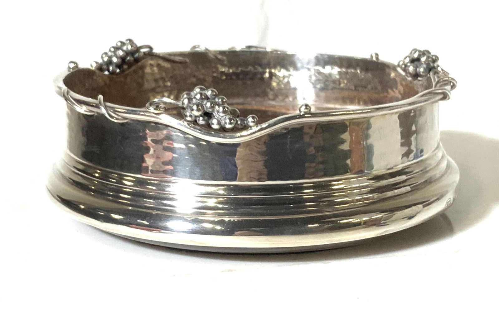 SJC Sterling Silver Grape Motif Bottle Coaster: Vintage Wine/ Champagne Bottle Holder/ coaster hammered metal gallery , grape bunched in relief around rim, and wood base. Bright and shiny with no evident tarnish. App 5 in. Diameter, 2 in ht. Marked