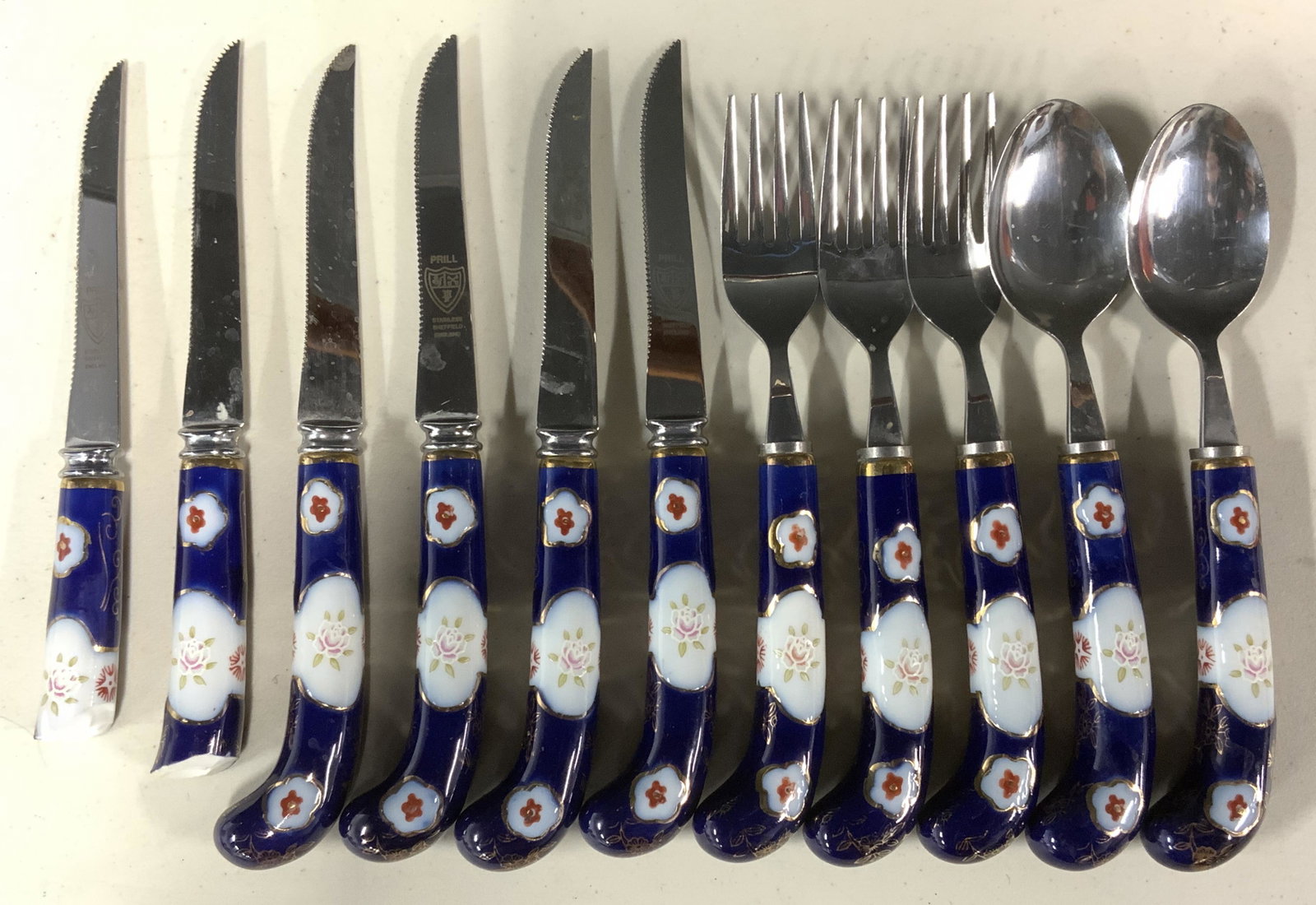 Group lot 11 PRILL Cutlery, England: Includes 11 ceramic handled utensils. 6 knives, 2 handles are broken, 3 forks, and 2 spoons. Knives read as PRILL Stainless Sheffield England, other utensils marked Stainless Steel. Surface wear prese