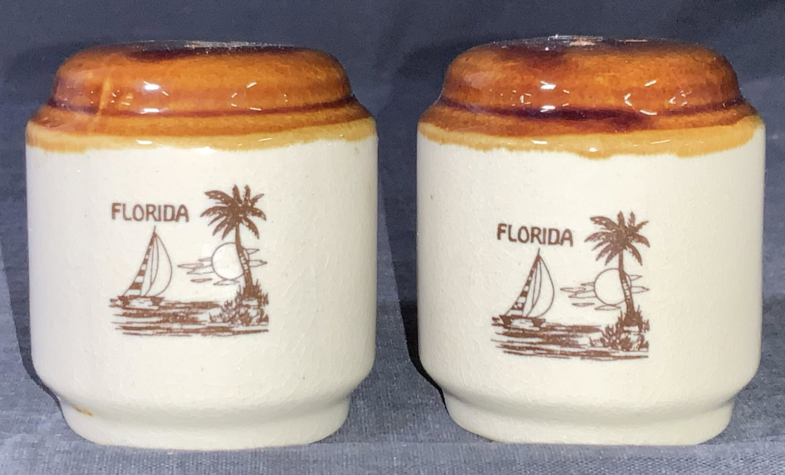 Set of Ceramic Florida Salt & Pepper Shakers: A pair of salt and pepper shakers made of ceramic, bodies are a cream tone with a caramel top coating with a scene of a sail boat in the ocean with a palm tree with Florida on the front. Both shakers 