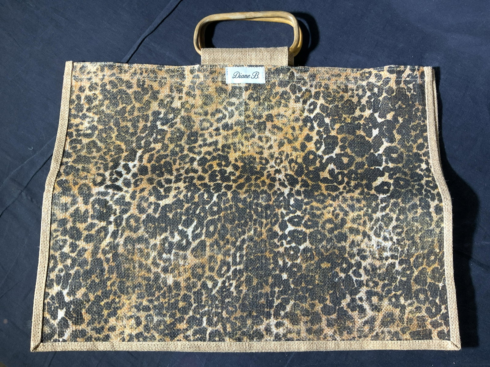 DIANE B. Animal Print Woven Wicker Bag: A woven bag made of wicker with a leopard print on the obverse side, tag on bag reads Diane B. And the handles of the bag are made of a light toned wood. The bag is approximately 21 x 6 in wide and 19