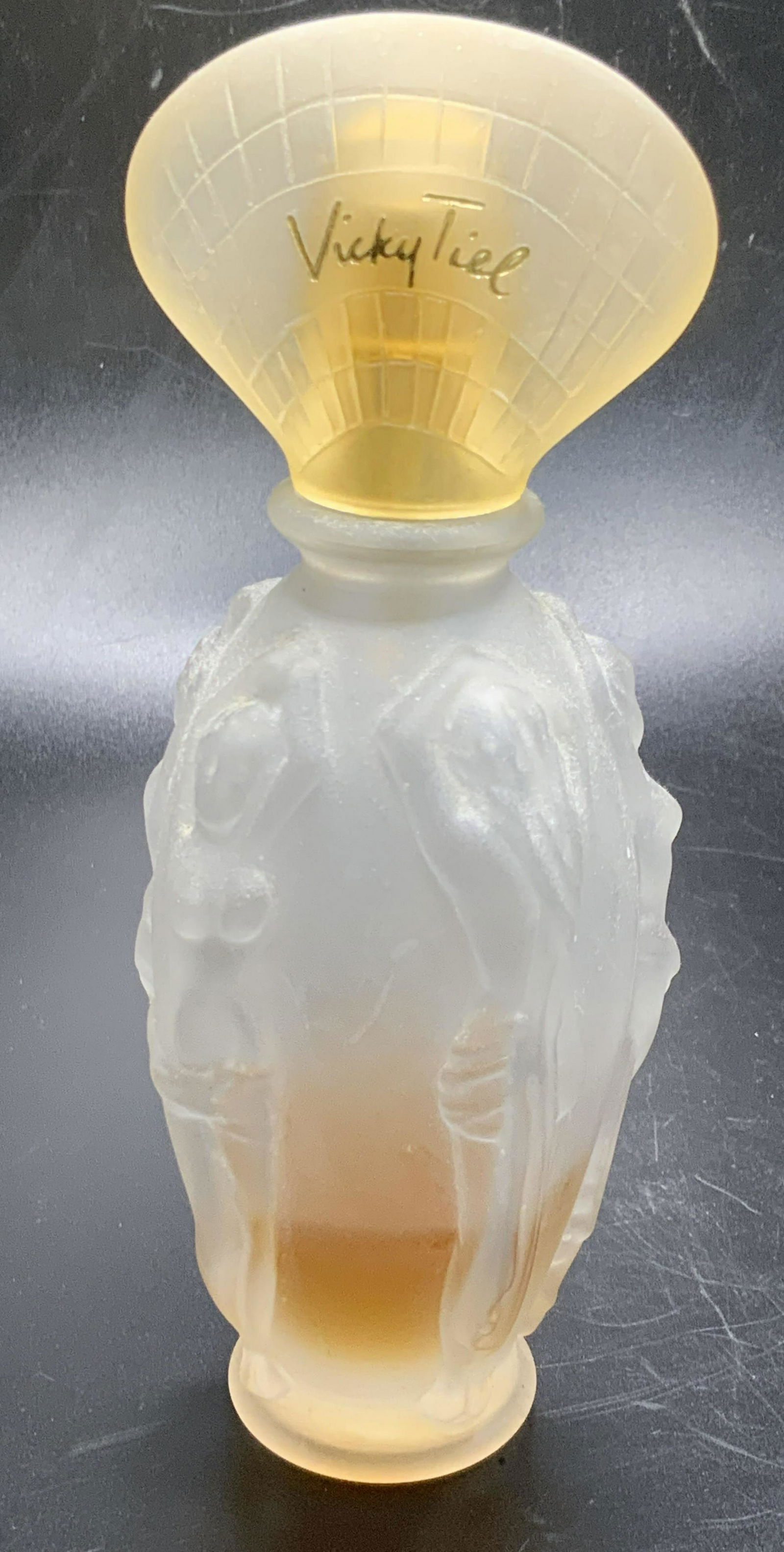VICKY TIEL Glass Nude Female Motif Perfume Bottle: Vintage frosted Glass Nude Female Motif Perfume Bottle. Women are raised, partially nude, arms reach up above their heads. Cap reads VICKY TIEL. Some liquid contained in bottle. Measures approx 6H x 2