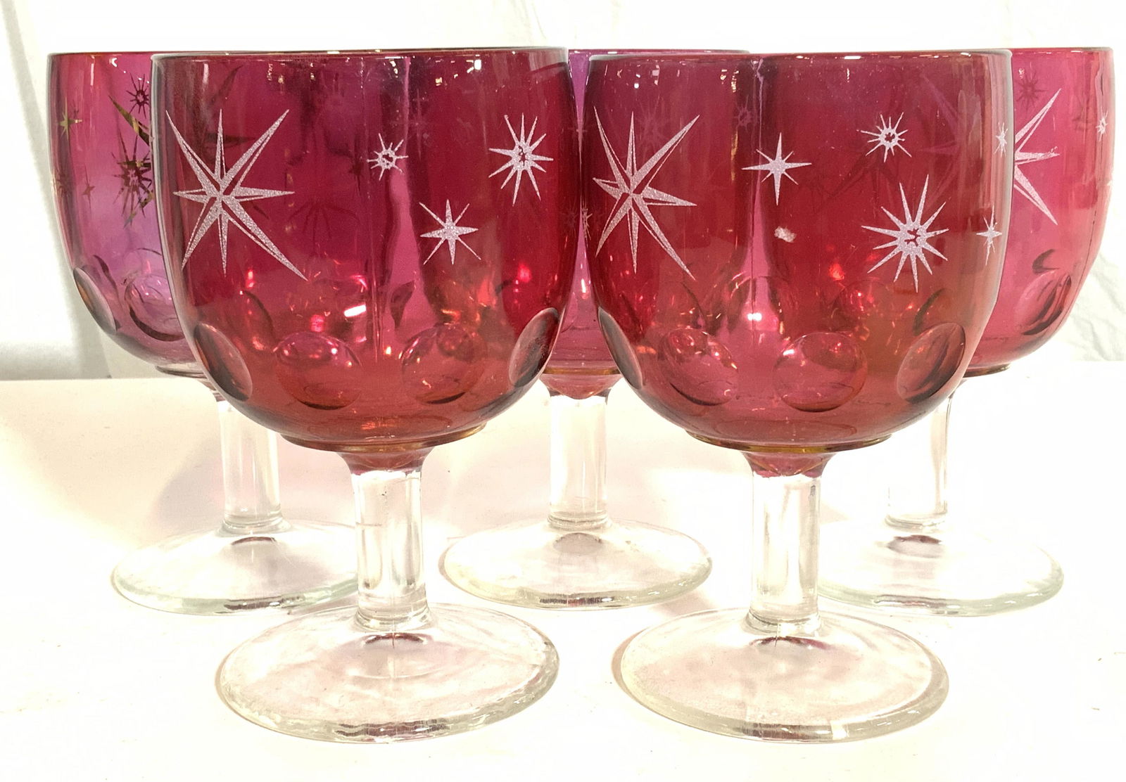 Set 5 Cranberry Glass Bohemian Glass Goblets: Lot includes 5 goblets. Each have cranberry toned basins and clear stems. Base of each stem is circular shaped. Most Basins have raised star shaped detail. Lower halves of each basin have circular sha