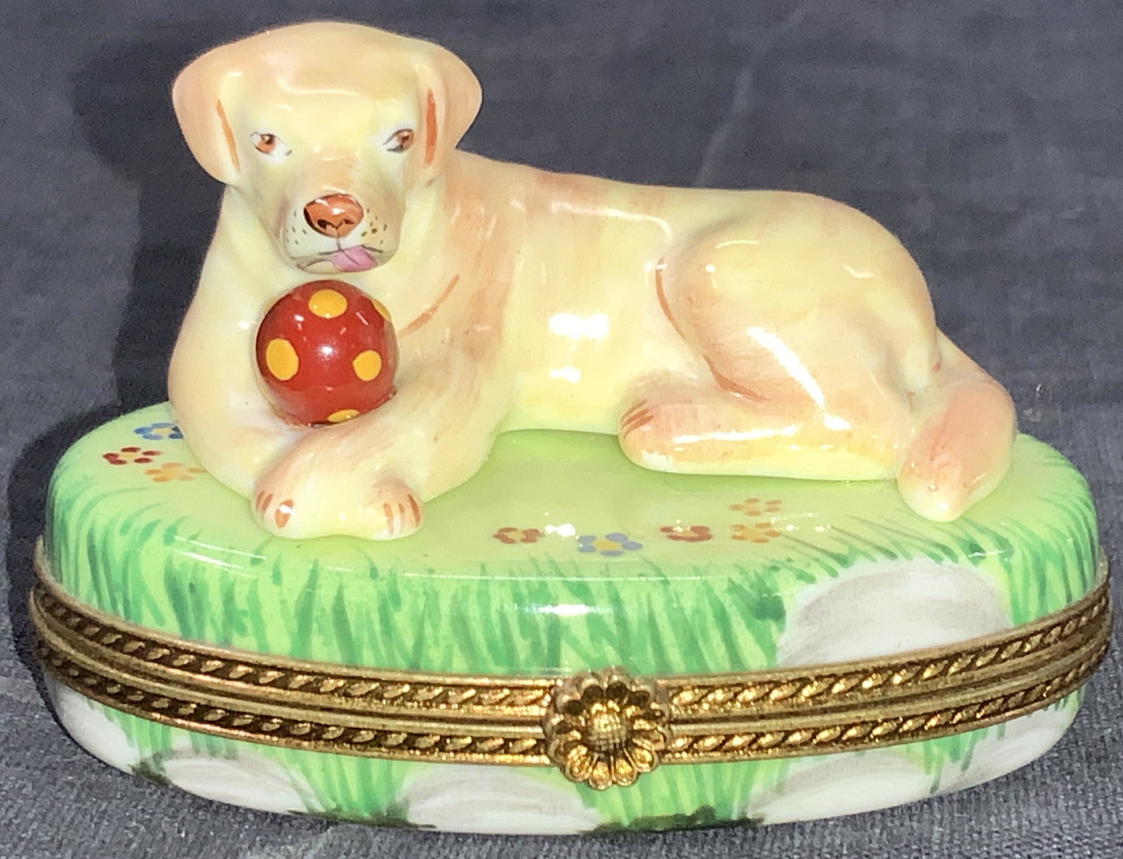 Limoges French Porcelain Golden Lab Pill Box: A porcelain trinket box made to resemble a golden lab with a ball laying on a grassy patch with flowers. Box has a lower compartment that opens to show a dog bone painted onto the bottom. Underside of