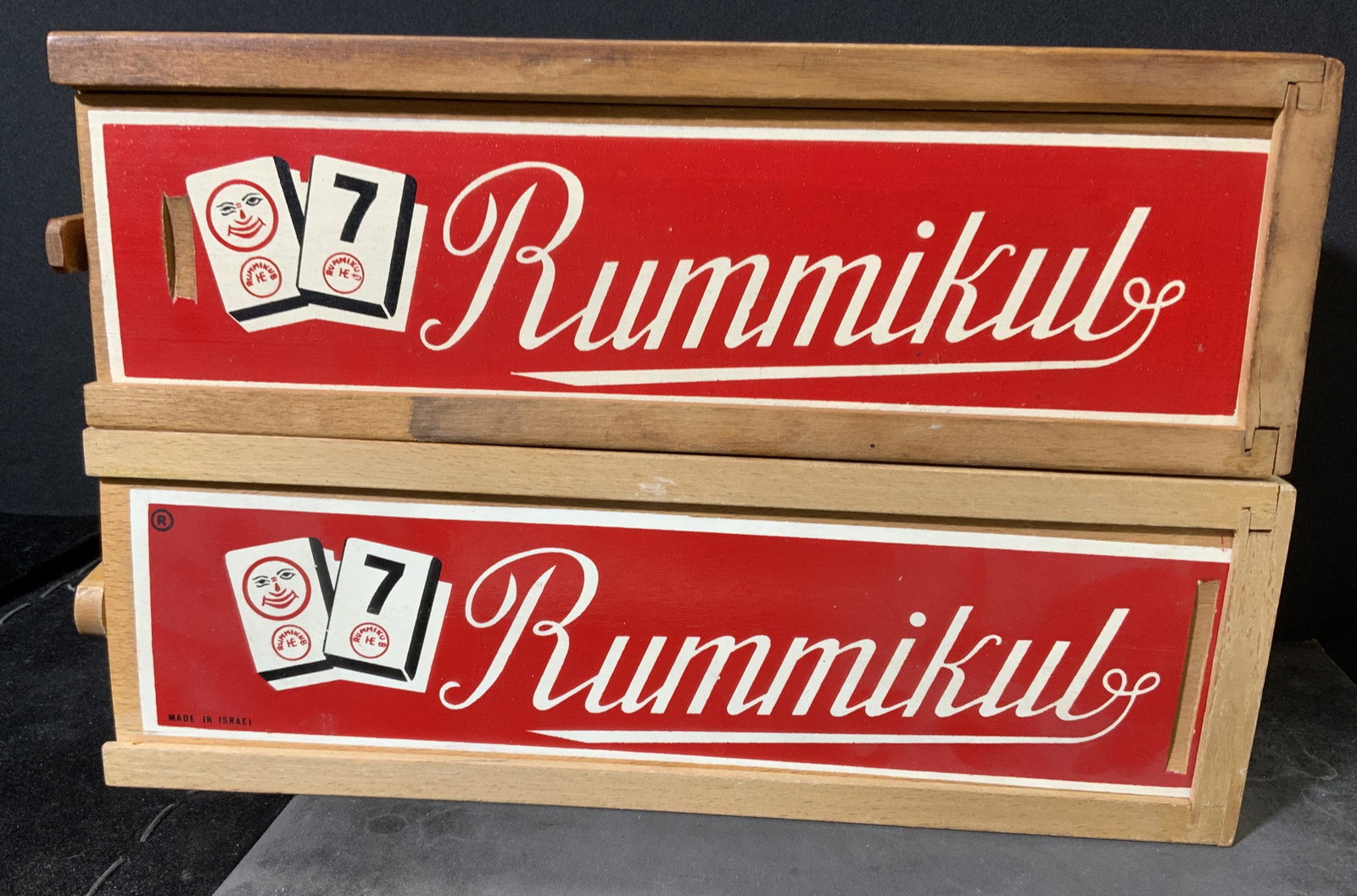 Lot 2 Rummikub Tile Games: Lot 2 boxes Rummikub. One box is missing several game tiles. Each includes 4 racks, rack fit into sleeve to form box. Splitting of wood and surface wear present. Largest box measures approx 2.5 inches