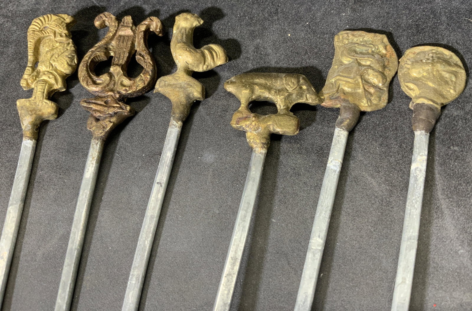 Group Lot 6 Vintage Brass & Steel Skewers: Includes 6 vintage skewers. Each is decorated with a cast brass ornament. 2 depict the faces of men, 1 rooster, 1 lyre, 1 pig, 1 depicts a man riding a kelpie. Surface wear and bending present. Each m