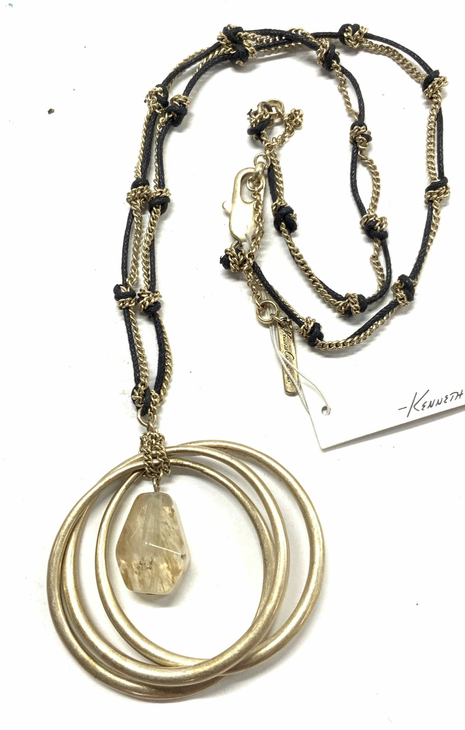 Kenneth Cole Signed Bohemian Style Necklace: Gold toned brushed metal necklace with cable chains with black cord. Pendant is made of three rings with a center Quartz style glass gem in the center. Marked on charm Kenneth Cole. Approximately 13 i