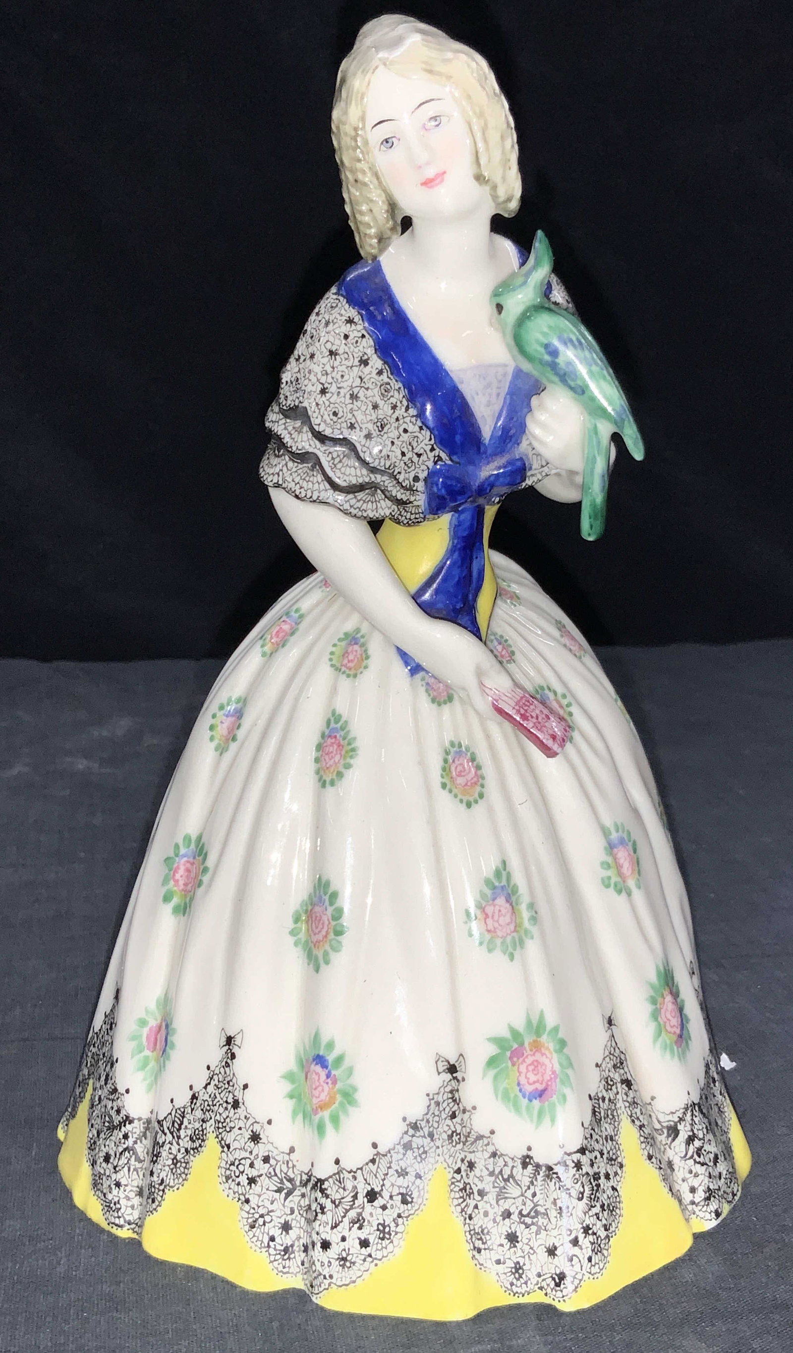 Vintage Hand Painted Porcelain Female Figure: A vintage female figure made of porcelain. Figure is adorn in 19th century dress, hand painted with yellow rims and intricate black lace style designs, pink and green flowers liter the dress with the 