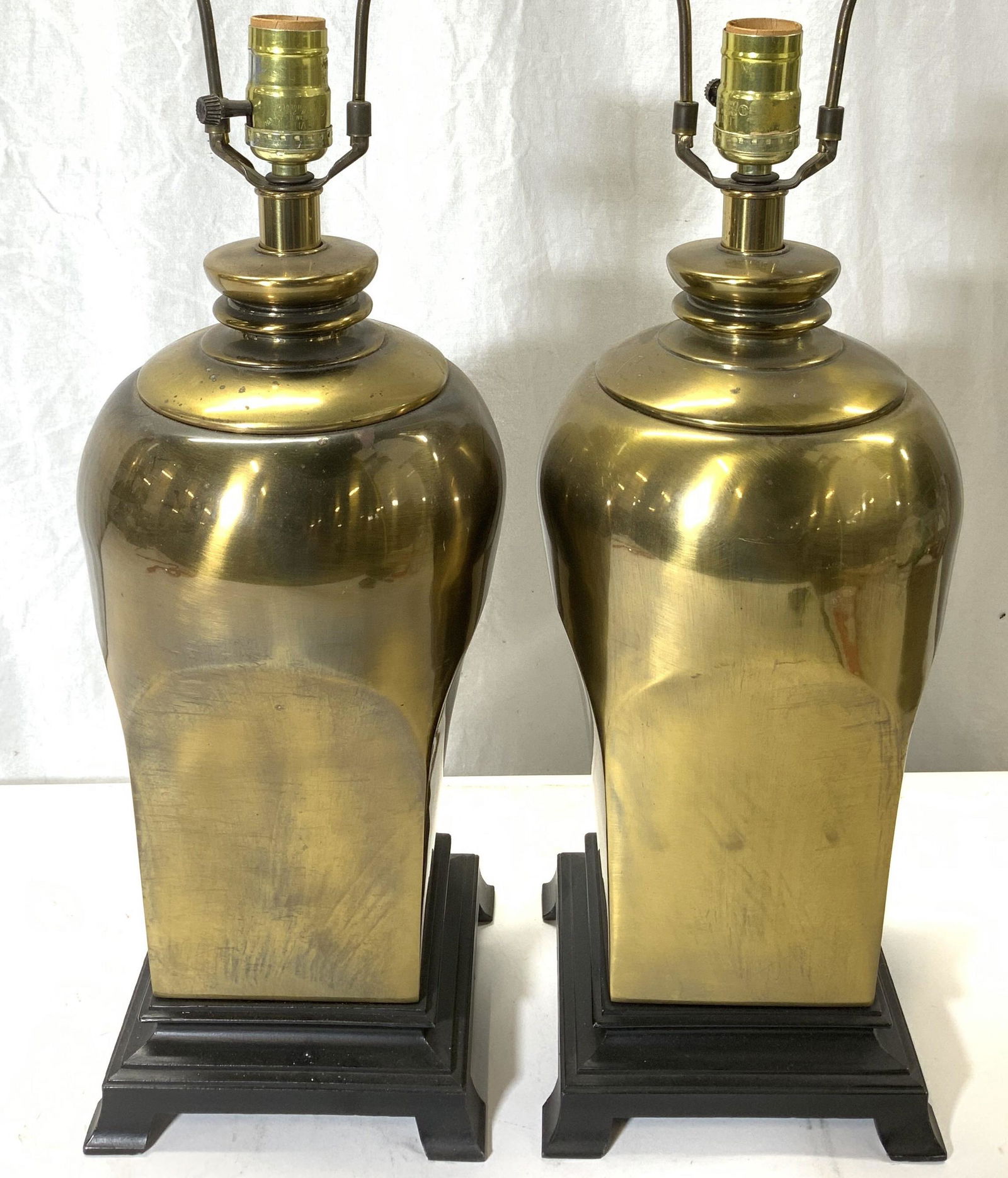 Pair Gold Toned Metal Lamps: Lot includes 2 lamps. Each are gold toned and metal possibly brass. Lamps each have black toned bases with feet. Each include harps & finials. Lamps measure 27 inches tall 7 inches wide. Lamps, metal