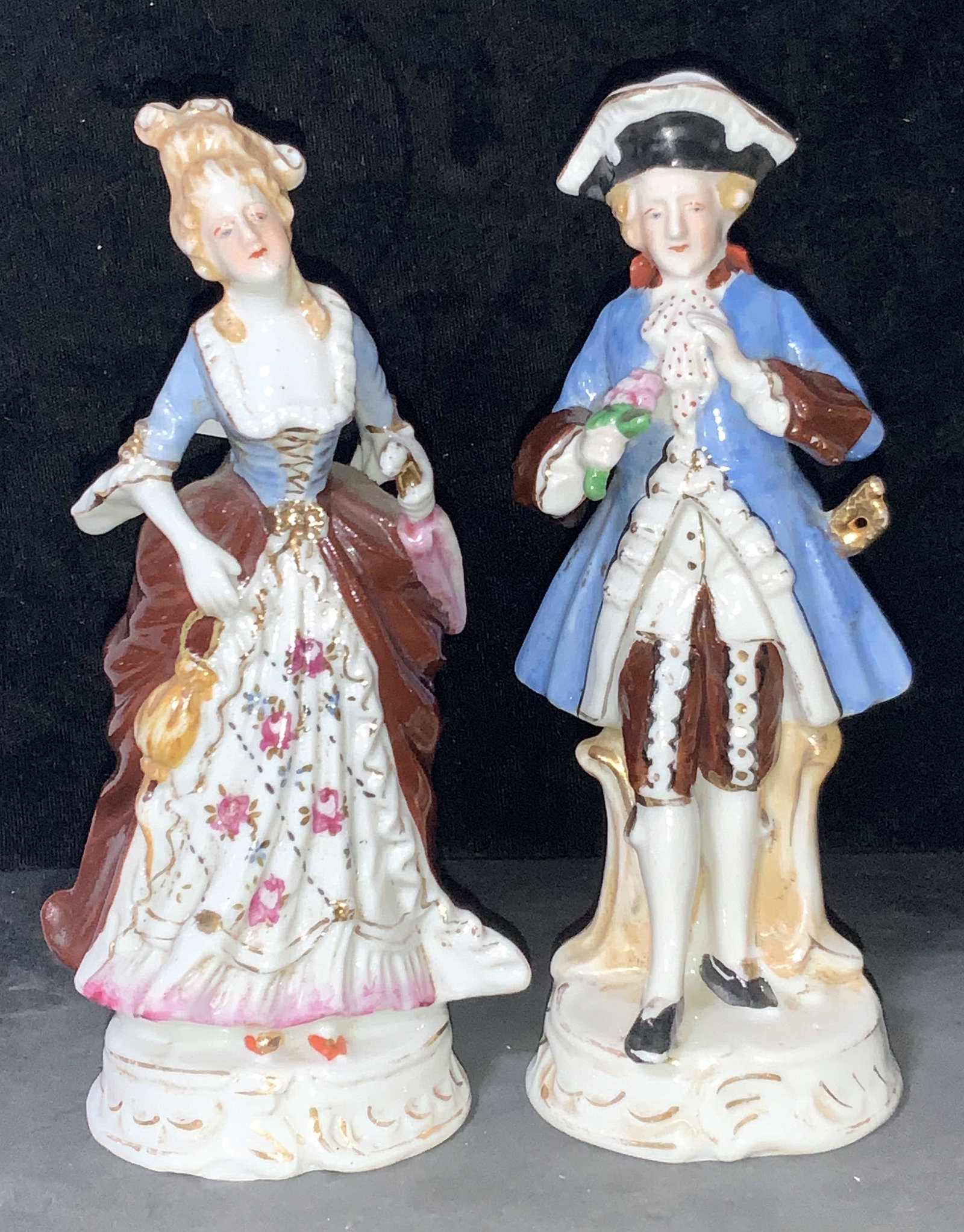 18th Century Style Porcelain Figures, Japan: A male and female porcelain figurines presented in 18th century clothing, hand painted. Man figure wears a black hat, blue coat and tights, the woman wears skirts of deep red, and floral motifs with a