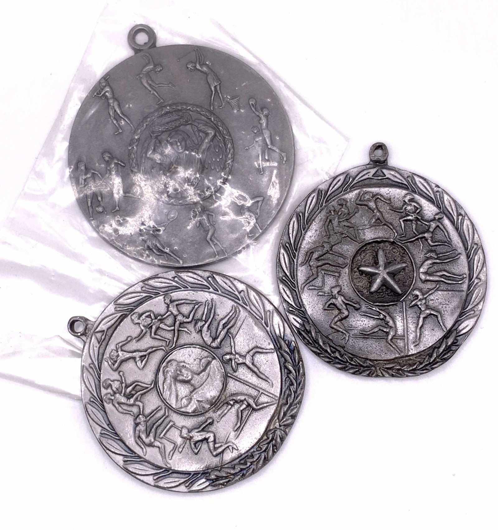 Lot 3 Vintage Sport Motif Metal Medals: Three vintage silver tone metal medals. The pieces depict people performing activities such as hurdling, running, tennis, and more. Raised metalwork designs. Bails to the top. Largest Measures approx 