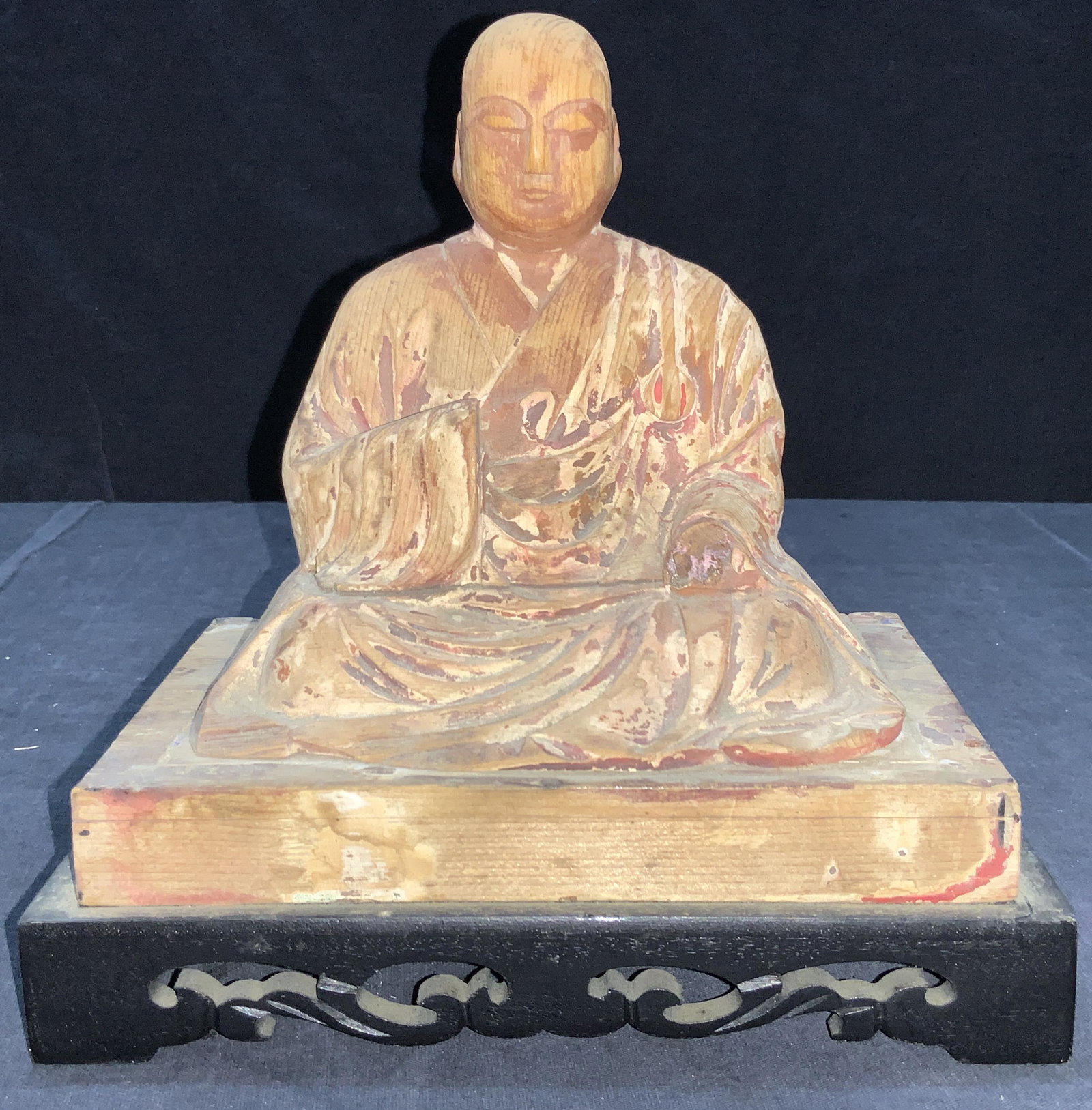 Carved Wooden Asian Monk Sculpture: A carved statute of a wooden seated figure, possibly a monk, in traditional robes affixed to a black carved wooden stand. Approximately 9 inches tall and 8.75 inches long and 5 inches wide. Statue has