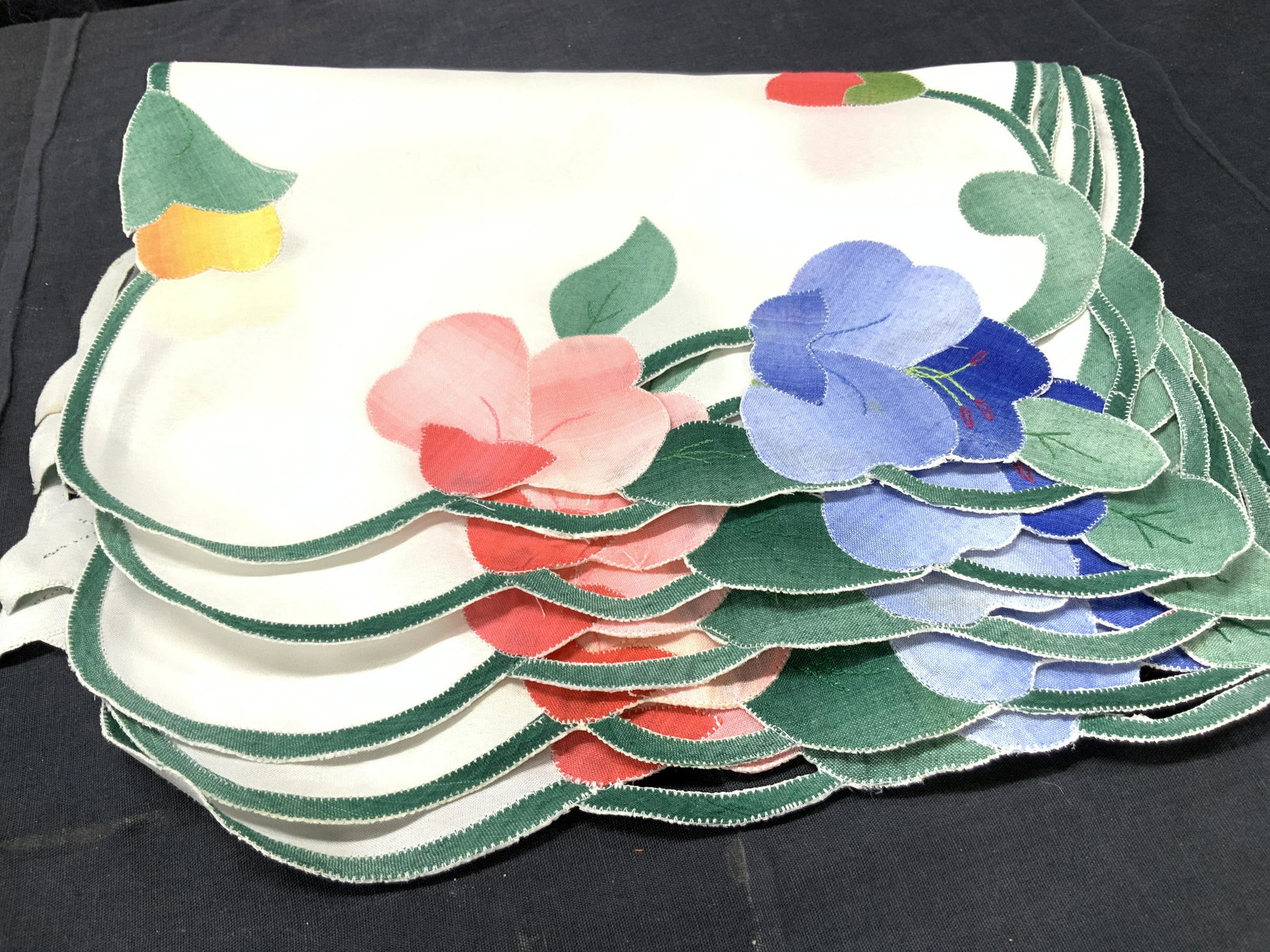 5 Vintage Floral Fabric Place Mats: 5 vintage place mats with floral appliques of blue, pink and yellows with green edges to resemble stems and leaves. Approximately 17.5 x 12 inches. Minor blemishes to certain mats. Fabric mats, tablet