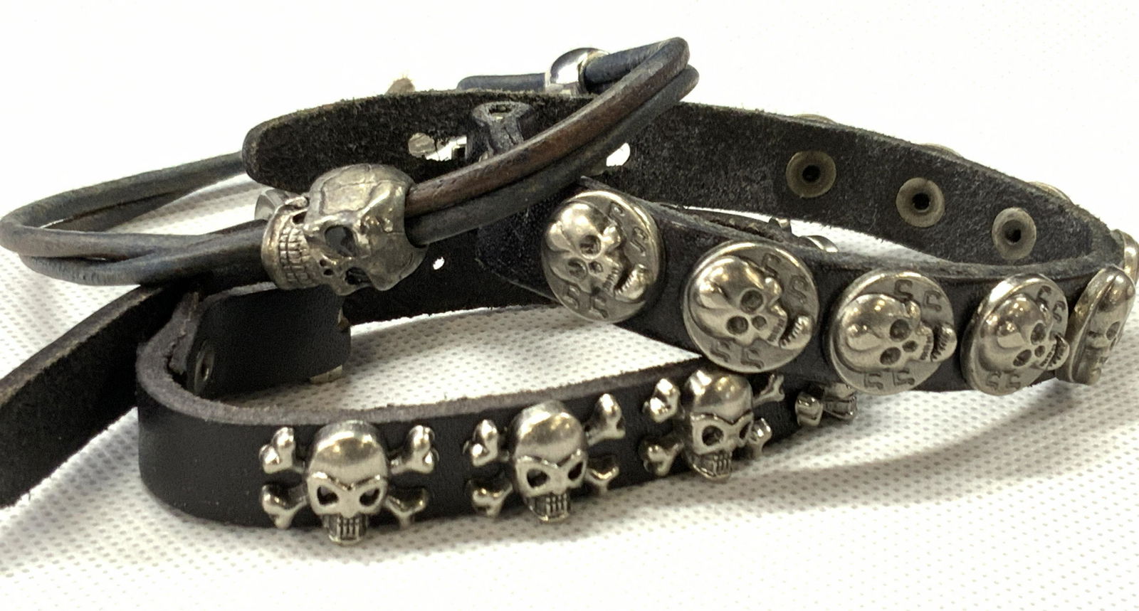 3 Vintage Leather Skull Bracelets: Group lot of 3 leather skull bracelets. 1 buckle bracelet with thick leather band with skull and cross bone charms affixed to the band. 1 hook bracelet with skull charms throughout. And 1 black corded
