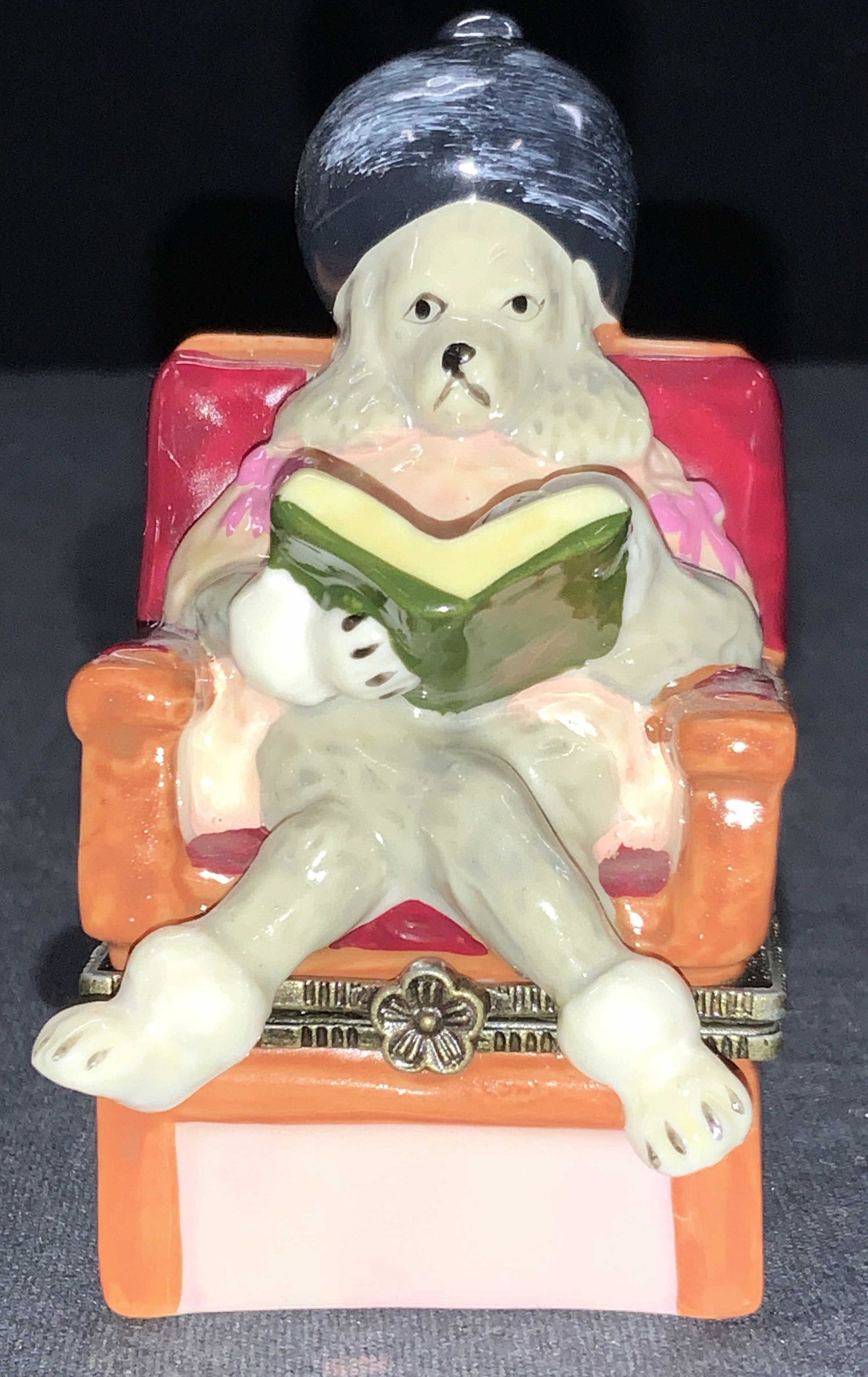 Porcelain Poodle Pill Box: Porcelain trinket box depicting a poodle in a salon chair reading a book. Chair opens near base with a miniature bone inside. Approximately 4 inches tall and 2 inches wide. Porcelain trinket box, pood