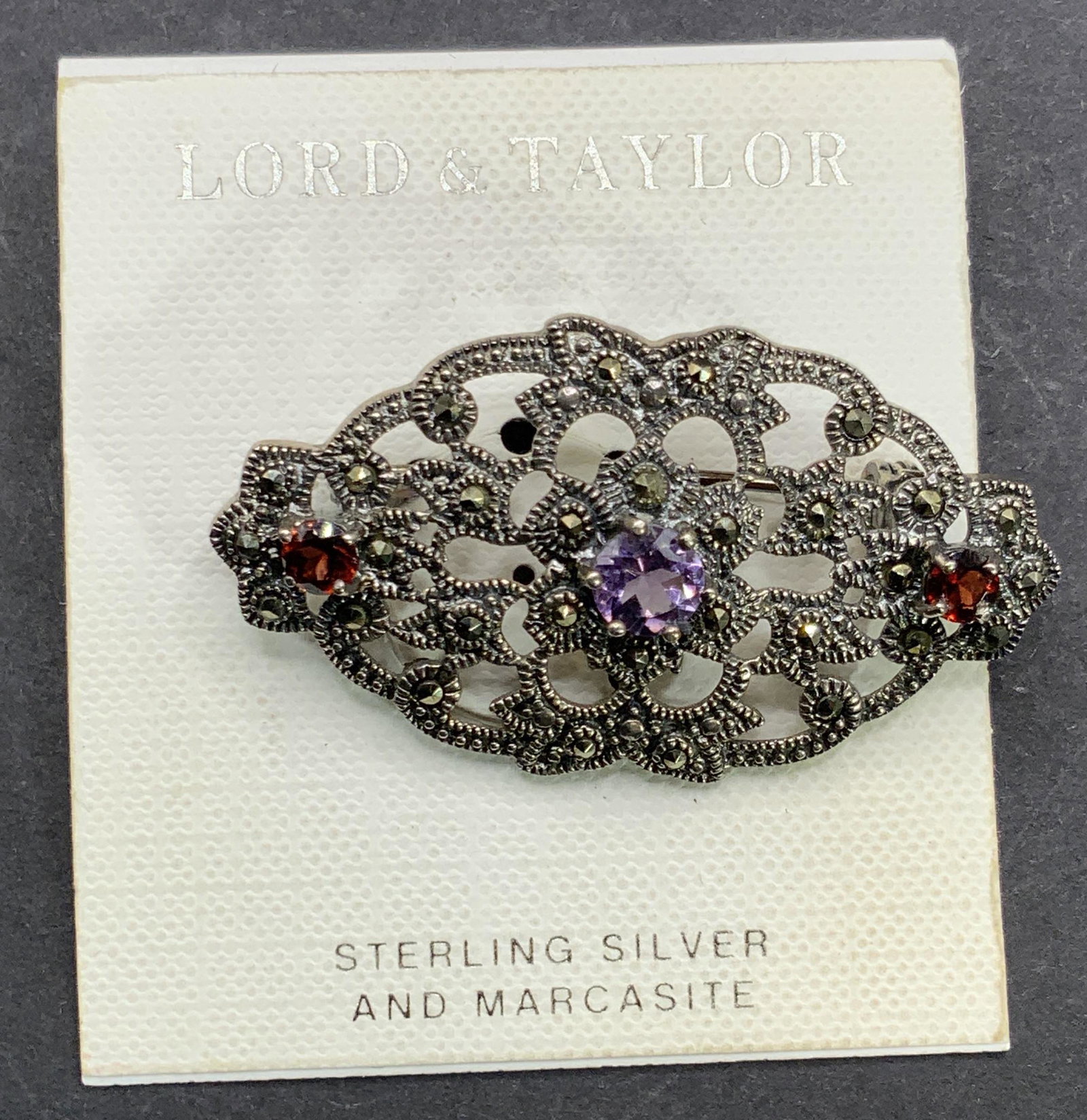 LORD & TAYLOR NWT Sterling Silver Marcasite Brooch: A sterling silver filigree brooch studded with marcasite. Three faceted stones to the center, one amethyst and two garnet, Measures approx 1L x 1.75W in. Total weight .36 ozt. Affixed to original pack