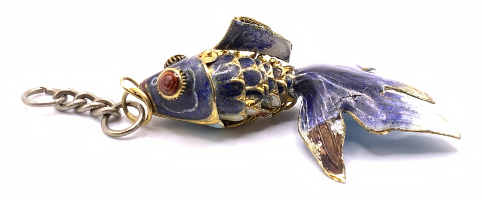 Vintage Articulated Cloisonne Fish Pendant: A vintage articulated fish form cloisonne pendant. Crafted from gold tone metal. Adorned with colorful blue and saffron hues. White accents. Bail to obverse. Silver tone metal chain attached to bail. 