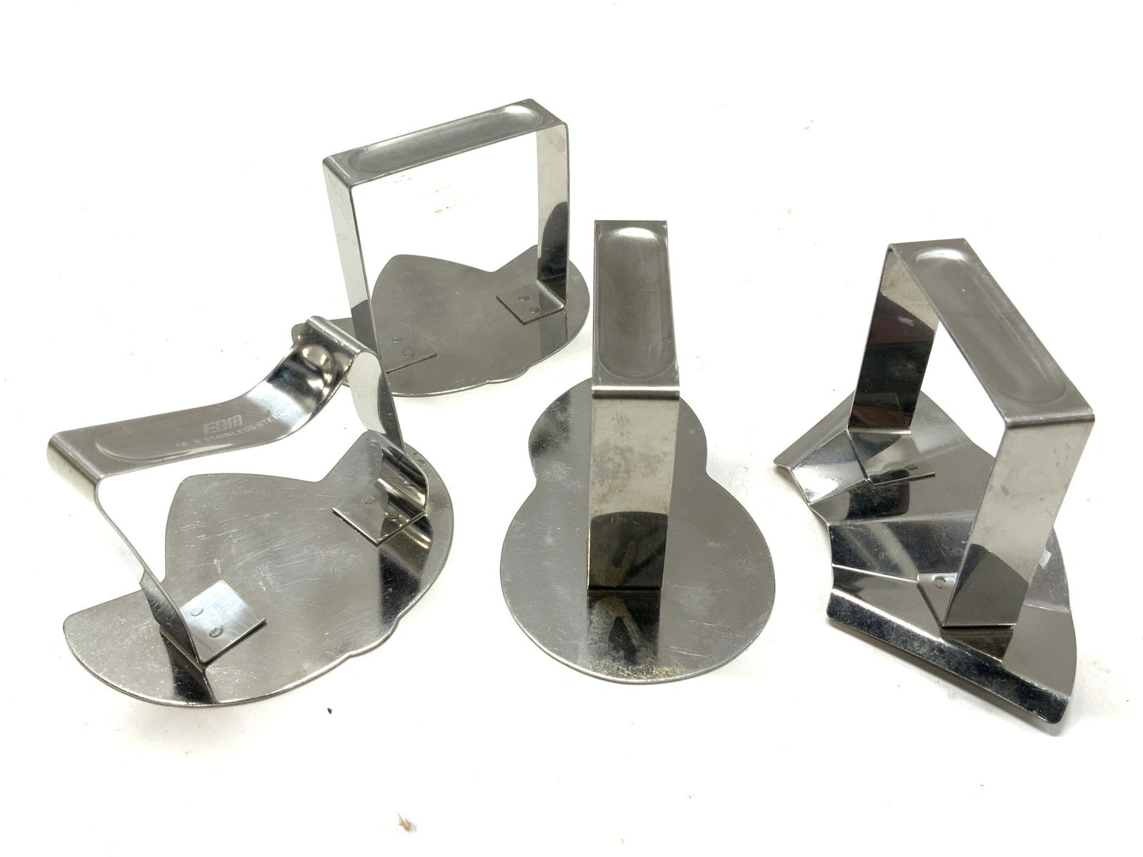 EBM Stainless Steel Shaped Napkin Holders: 4 stainless steel napkin holders with bases in varying shapes. 2 with the shape of leaves or buck feet. 1 with a folded steel base and 1 with a gourd shape base. Marked on handle, EBM Stainless Steel.