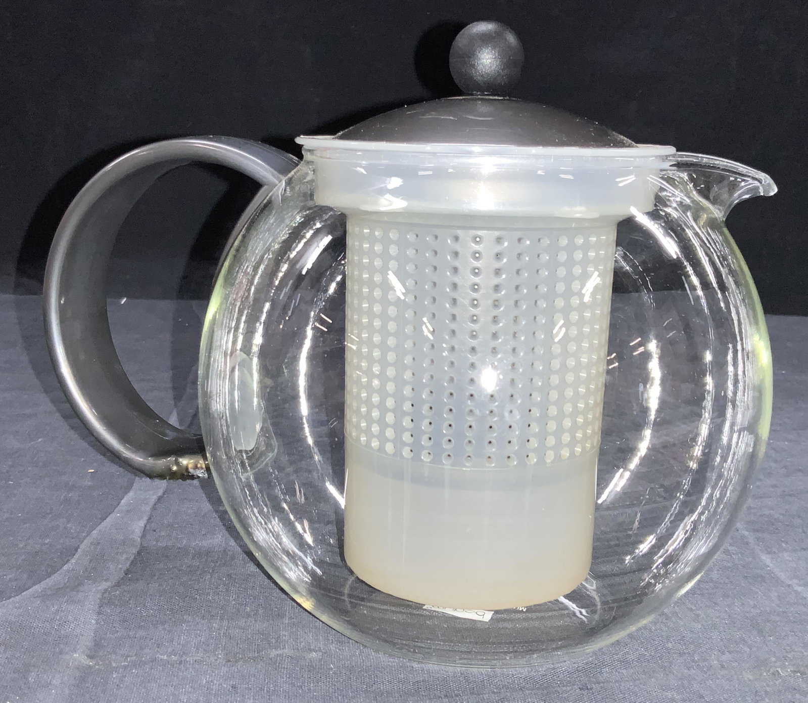 BODRUM Clear Glass Tea Kettle W Diffuser: A clear glass tea kettle with composite gray toned handle. Comes with tea herb diffuser with presser. Marked Bodum on underside. Approximately 7.25 inches tall. Burn marks to underside of handle. Prop