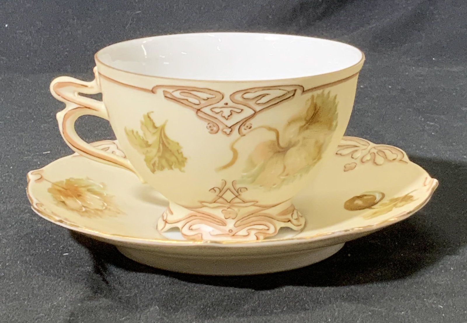 HERMANN OHME Silesia Porcelain Teacup & Saucer: Hermann Ohme Silesia Old Ivory floral motif and ornate flourish hand painted teacup and saucer in hues of ivory, beige, soft pink, and muted green. Signed SILESIA J4 on underside of teacup and SILESIA