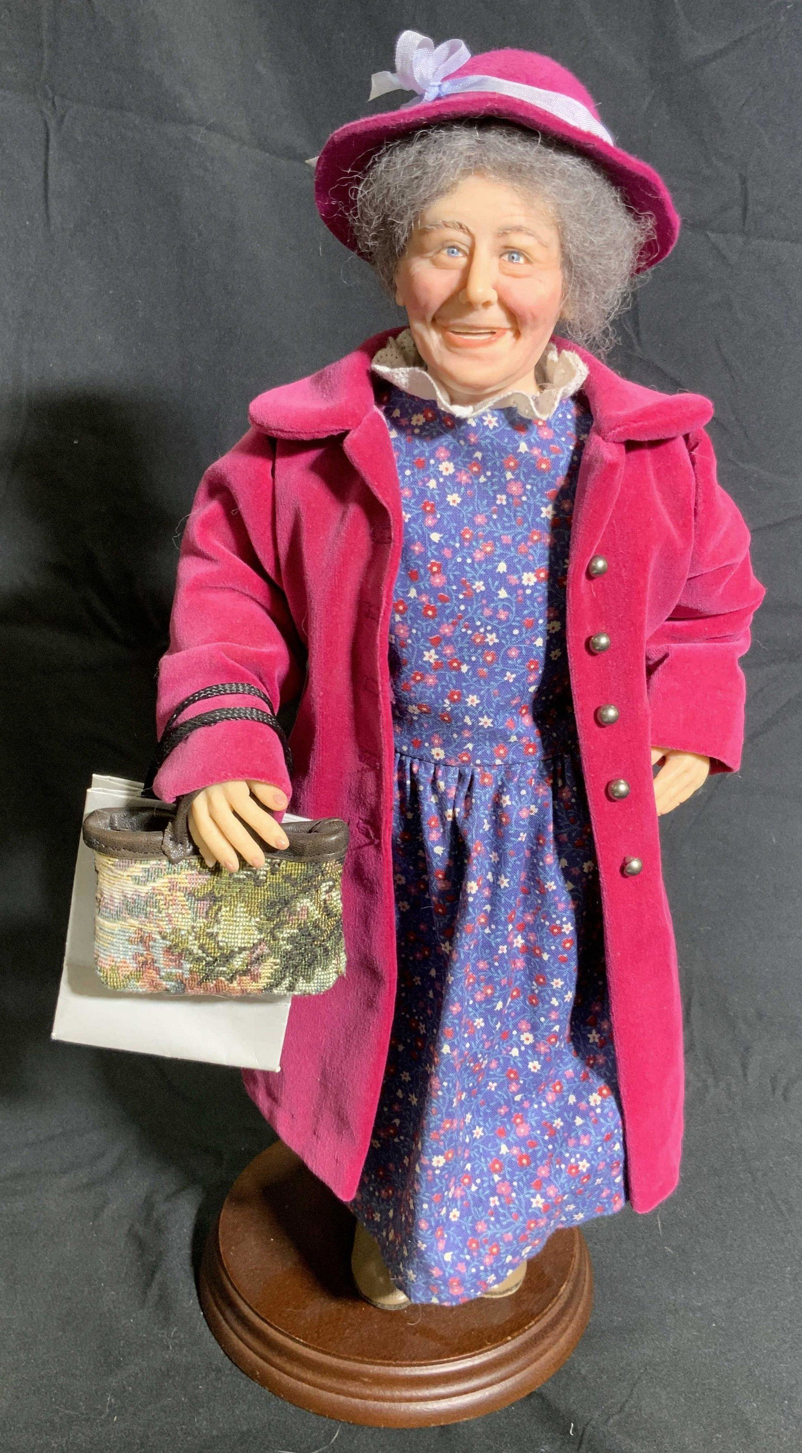 SARAH F. HAYNES Signed Elderly Woman Standing Doll: Subject is a elderly woman standing doll. She is attached to a wooden circular base. Appears to be crafted from a resinous material. Hand painted. She wears a floral dress and a pink coat & hat. She h