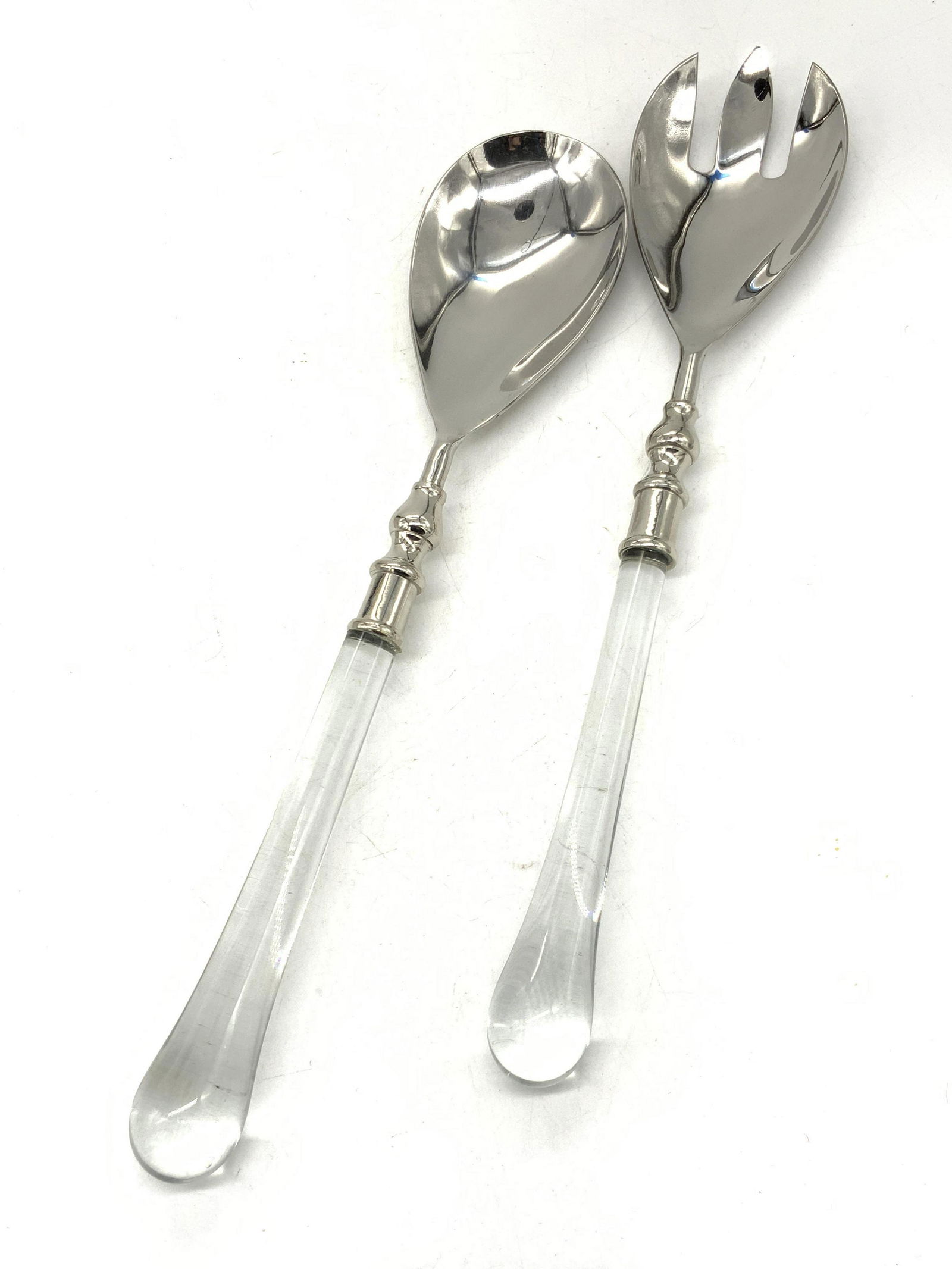 2 Clear Glass Handled Serving Utensils: Pair of serving utensils, white metal heads, possibly steel, with clear glass handles. 1 serving spoon, and 1 salad spoon. Approximately 13.25 inches long. Evident scratching to metal. Salad serving s