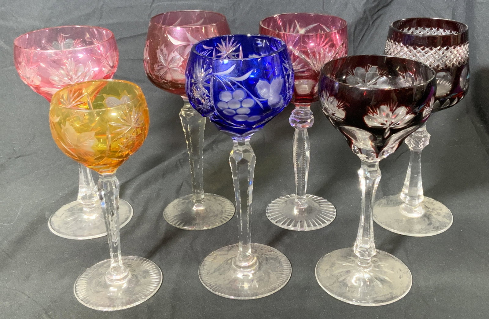 Group Lot Colored Floral Wine Glasses: Assorted group of 7 colored wine glasses in varying styles. Many with floral or vegetal motif. Glasses in hues of marigold, blue, and maroon. Sizes vary, largest measures approx. 8 inches tall and 3.5