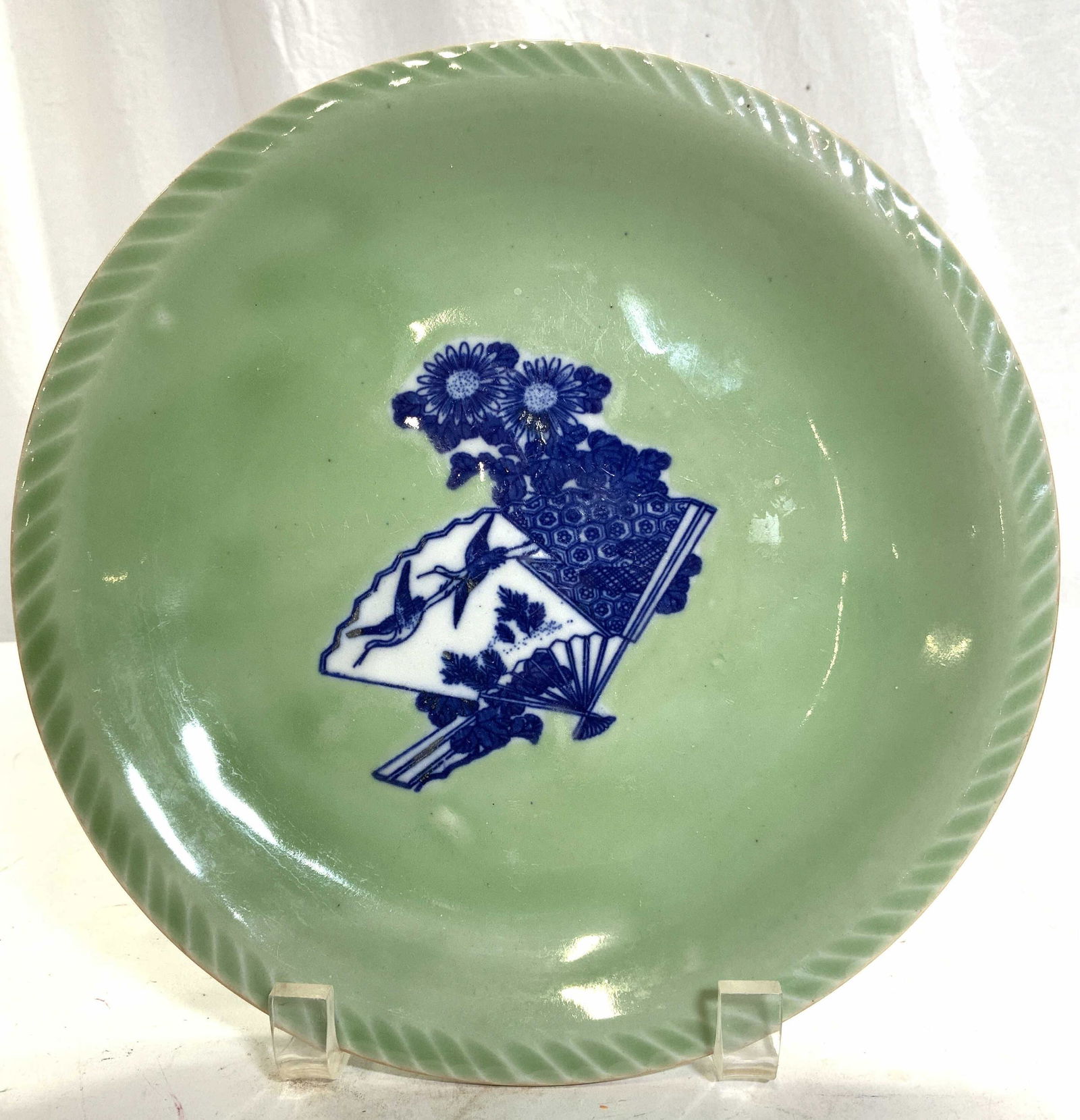 Vintage Asian Stoneware Celadon Plate: Plate is vintage possibly antique. Piece is thick porcelain. Plate is celadon toned with blue & white floral, leaf, crane & hand fan detail. Rim has relief detail. Plate measures approx 9.25 inches wi