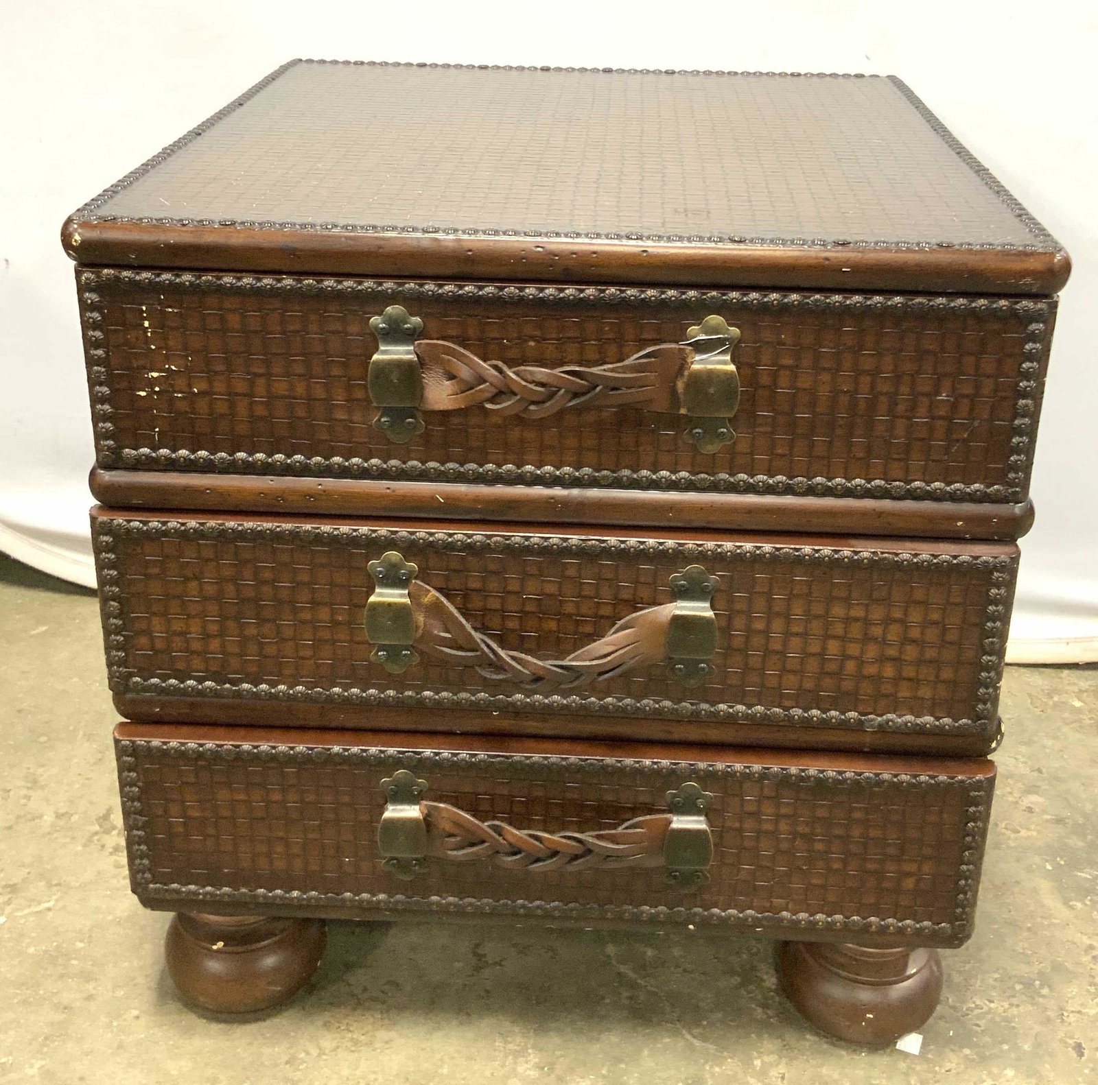 Vintage Etched Wooden Three Drawer Nightstand: A rich brown hued wooden nightstand. Square pattern etched surface. Floral stud detail. Three drawers with leather style drawer pulls and metal hardware. Four carved feet. Drawer pulls need addressing