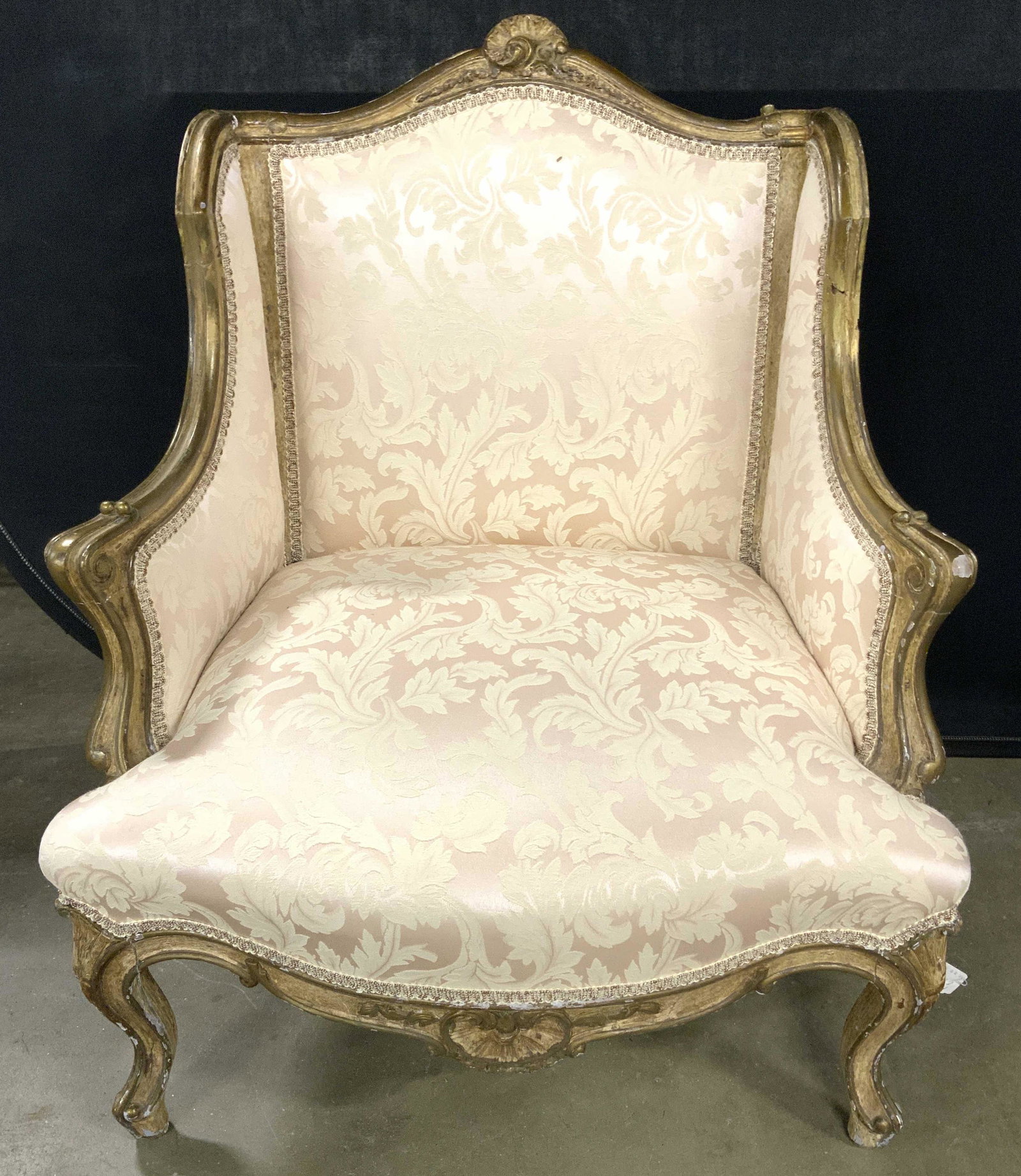 Antique French Louis XV Style Low Winged Armchair: Armchair is vintage possibly antique. Piece has winged backrest & gilt wood frame. Armchair has upholstery with gimp edges. Piece has ornately carved feet. Piece measures approx 32 inches tall 26 inch