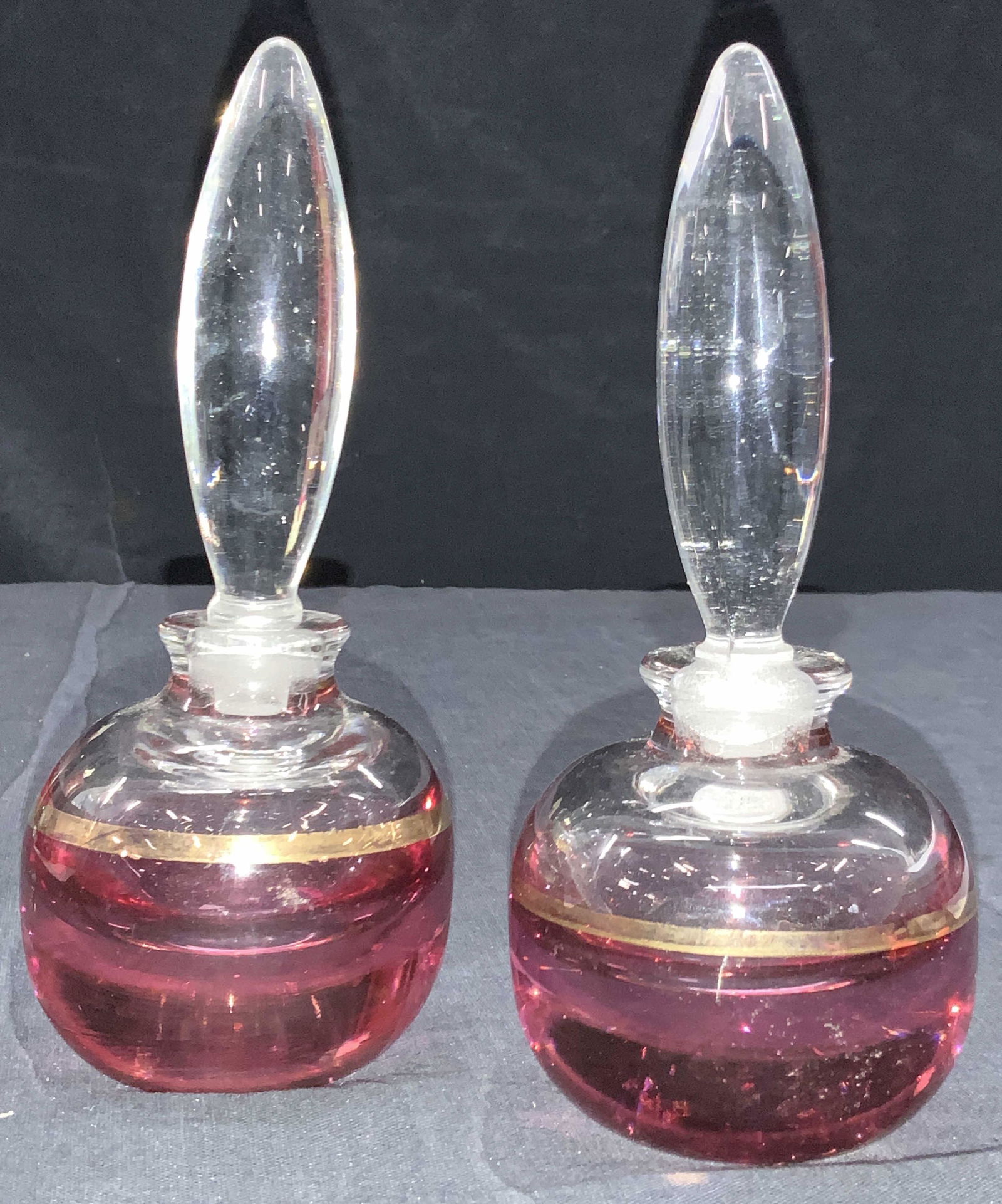 2 Gold Gilt Art Glass Perfume Bottles: Pair of 2 glass decanters with rounded bodies on a cranberry tone with gold gilded bands. Stoppers are long in shape. Approximately 6.25 inches tall. Evident wearing to gold gilt. Art glass perfume bo