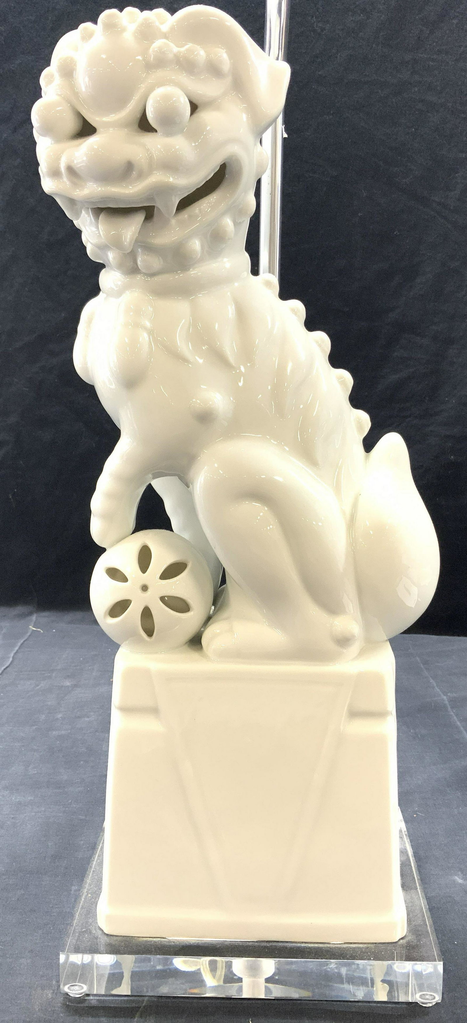 Blanc De Chine Porcelain Temple Lion Lamp: Lamp is white toned and porcelain. Piece is in shape of a temple lion with paw on spherical object. Base has rectangular prism shape and appears to be acrylic. Total height 30 1/4 in tall. Base and fo