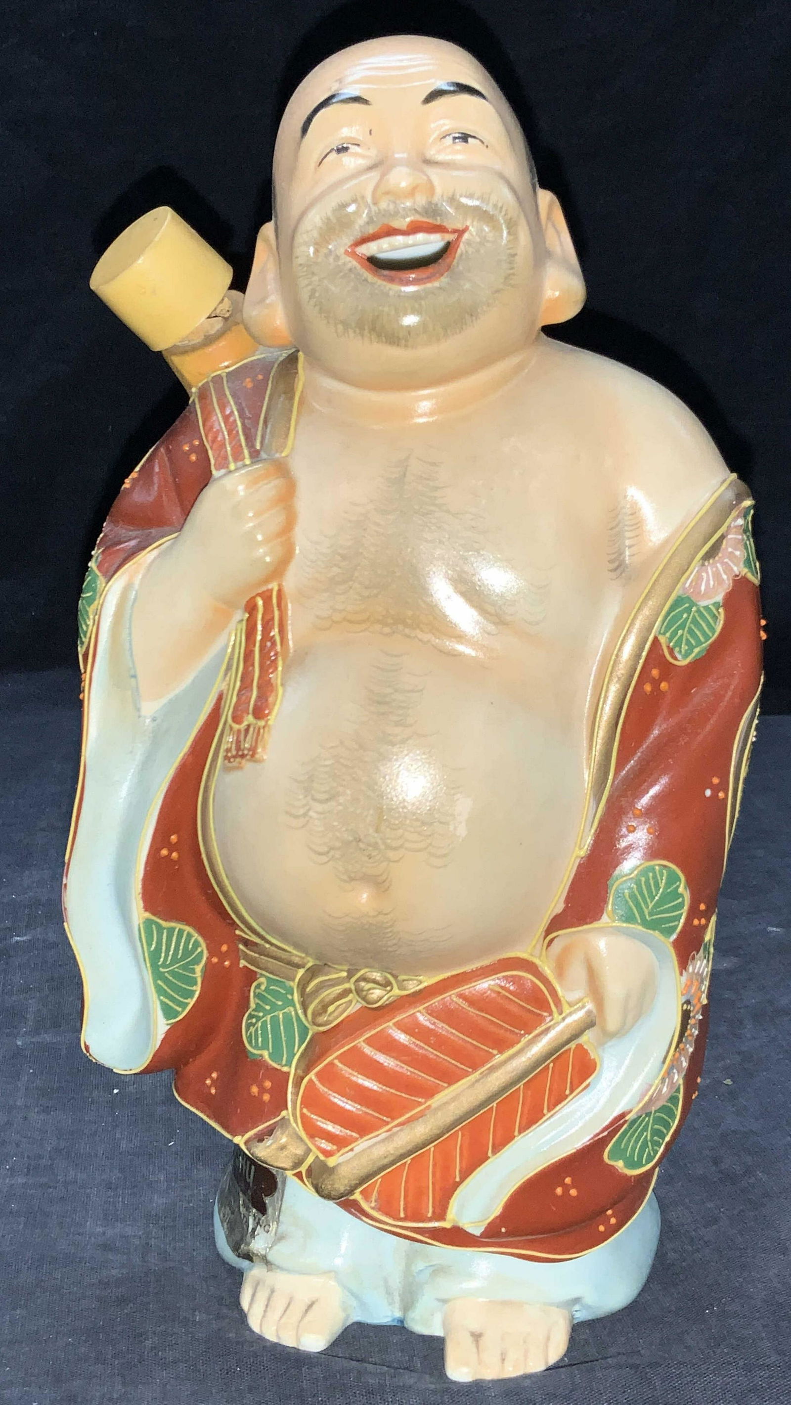 HOUSE OF KOSHU Hand Painted Monk Sake Bottle: A hand painted ceramic figure of a monk, painted in the Satsuma style with red toned robes with raised gold painted outlines. Monk is holding fan in one hand and a strap for the gourd on the back. Gou
