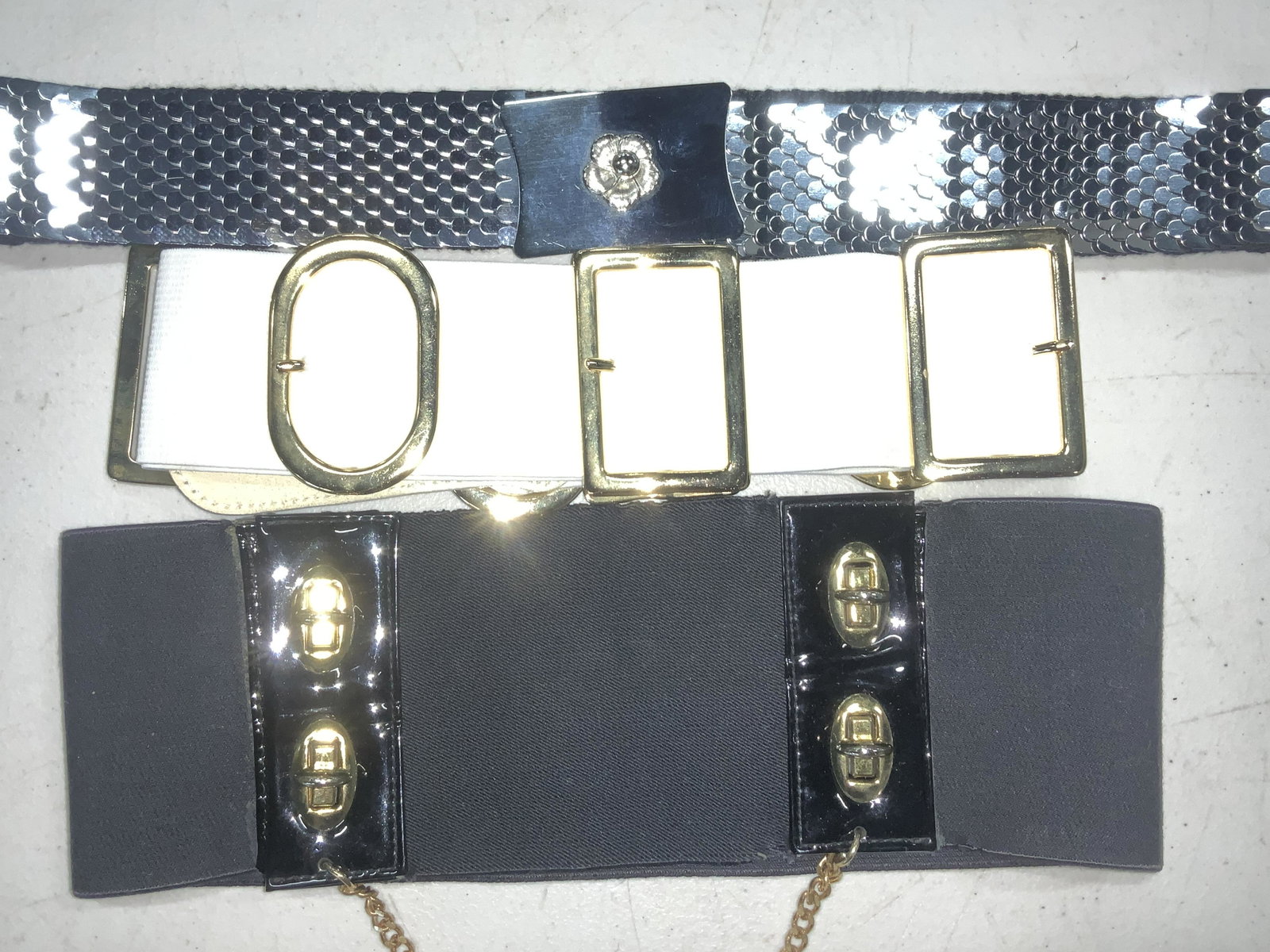Grp 3 Vintage Stretch Belts, c. 1980s: 3 distinct vintage Ladies belts, all stretch. One gunmetal metallic fish scale style belt, hook and loop latch, buckle is polished silver tone metal with center flower in relief. 30.5 in. L White Stre