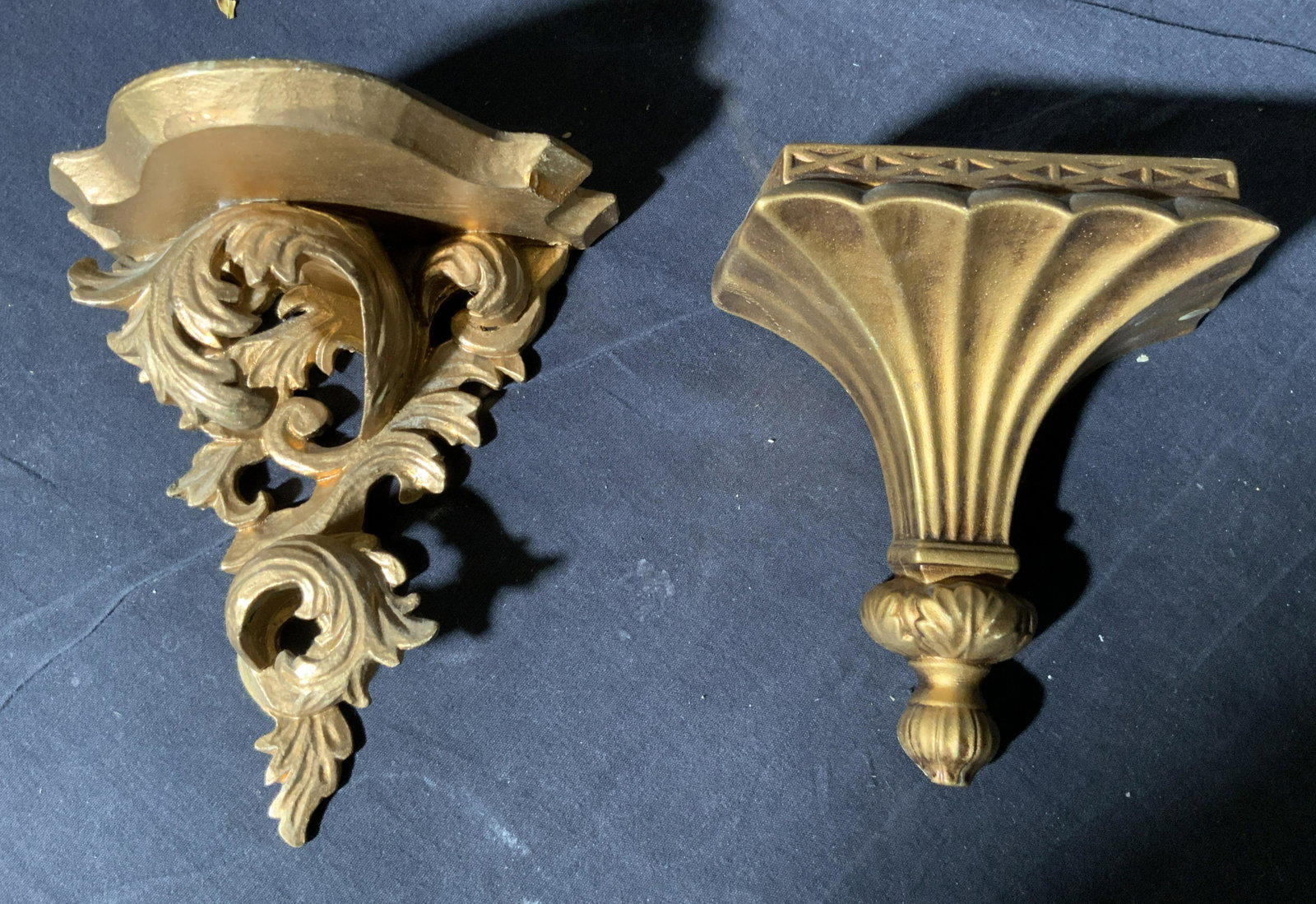Lot 2 Gilt Plaster & Giltwood Wall Shelves: Lot includes 1 gilt wood wall shelf and 1 gilt plaster wall shelf. Gilt wood wall shelf has ornate scroll and leaf structures. Gilt Plaster wall shelf has x shaped relief detail near top. Lower end of