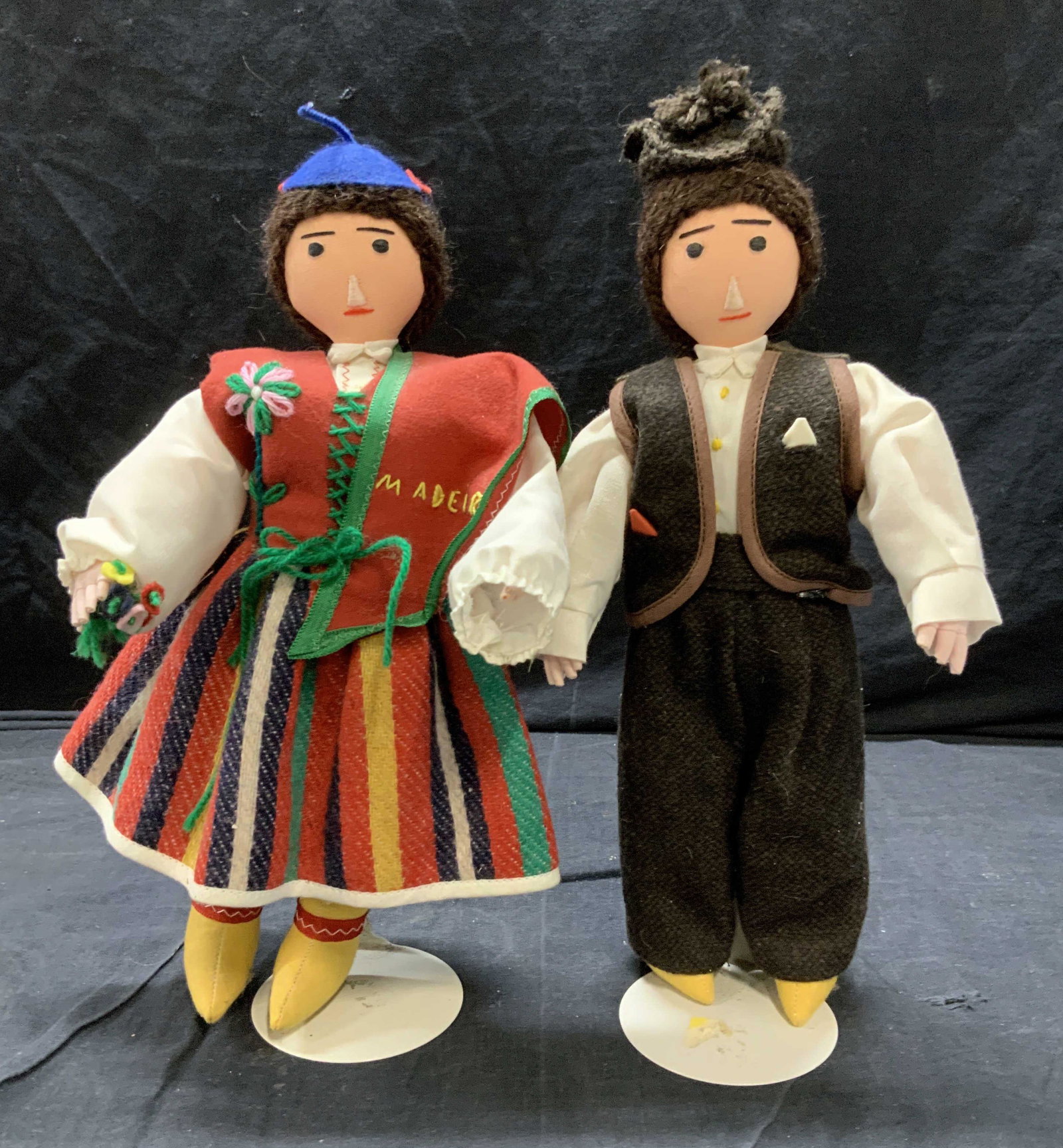 Pair Portuguese Folk Art Dolls on Stands: Artisanal folk Art dolls, one lady and one male, wearing cultural Portuguese outfits, lady has a vest that reads Madeira. clothing accents with yarn embroidery and Fabric faces. Each measures 12 x 6.5
