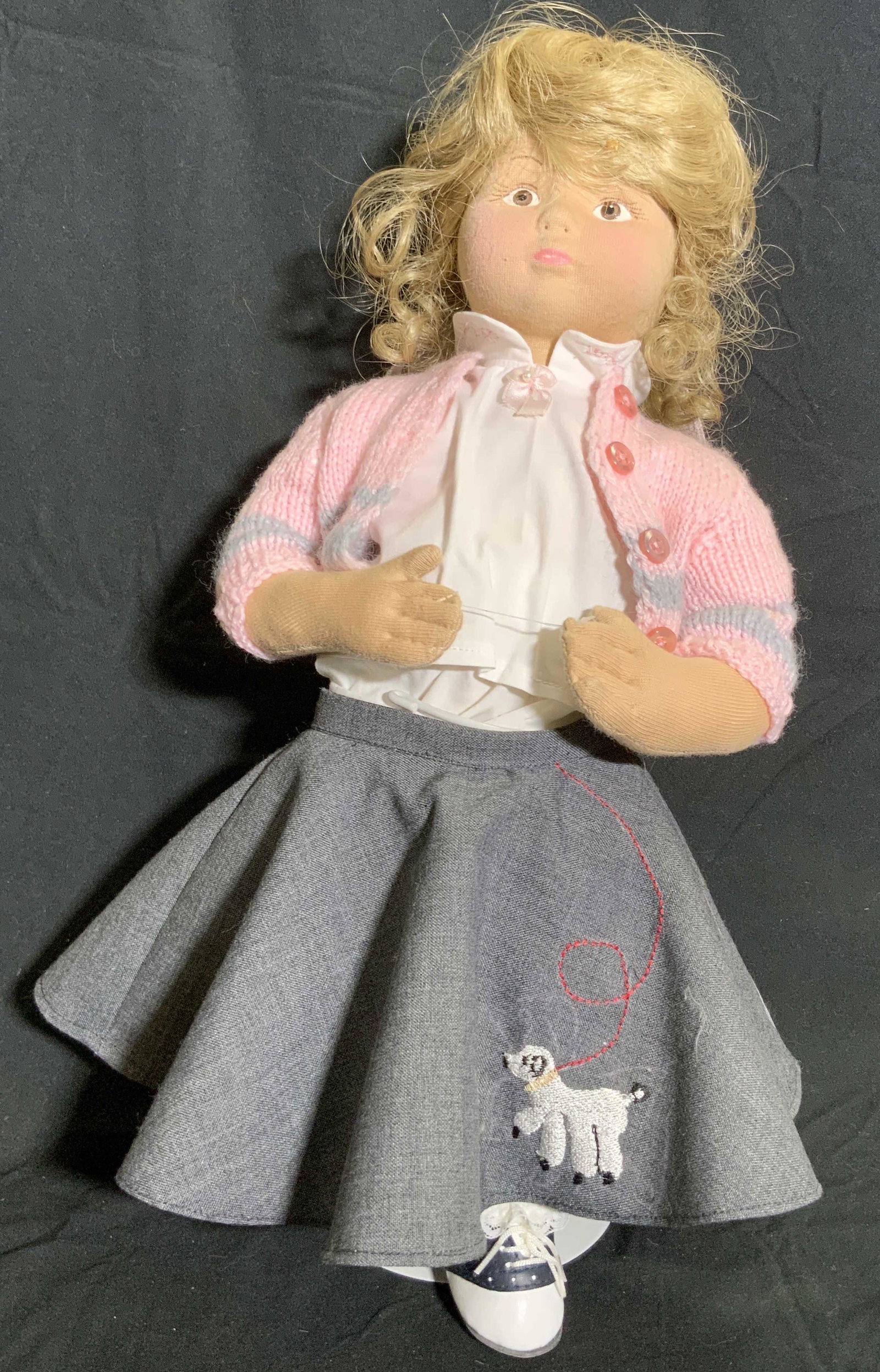 Vintage Fabric Doll W/ Poodle Skirt, Stand: A vintage fabric doll wearing a pink cardigan and gray embroidered poodle skirt. Hand painted details to face. Comes with a metal stand. Measured in stand. Measures approx 18H x 8W in. Some wear consi