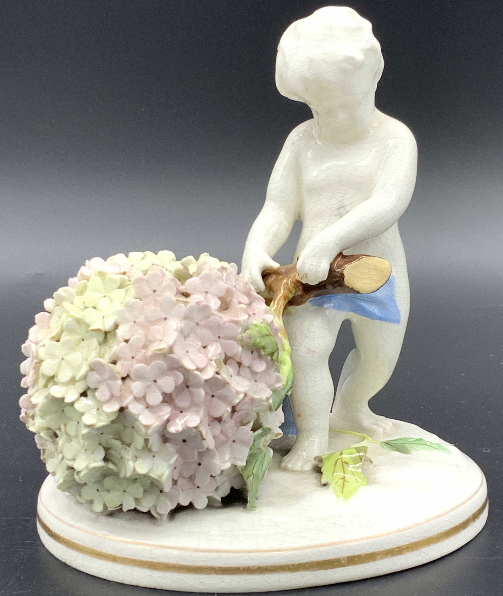 Porcelain Cherub Figural with Flower Bush: Hand painted and glazed porcelain figural/ statuette of Cherub putti holding onto a Branch. Unmarked vintage porcelain with glaze crazing. hand painted and hand formed individual flowers. Measures 5.7